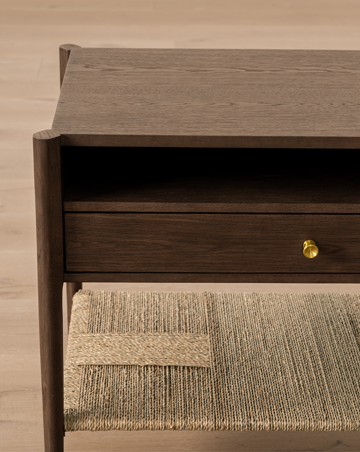 The Dana Walnut Oak Nightstand features a single drawer with a round brass knob, an open shelf above, and a woven rattan lower shelf, all set on a light wood floor, by McGee & Co.