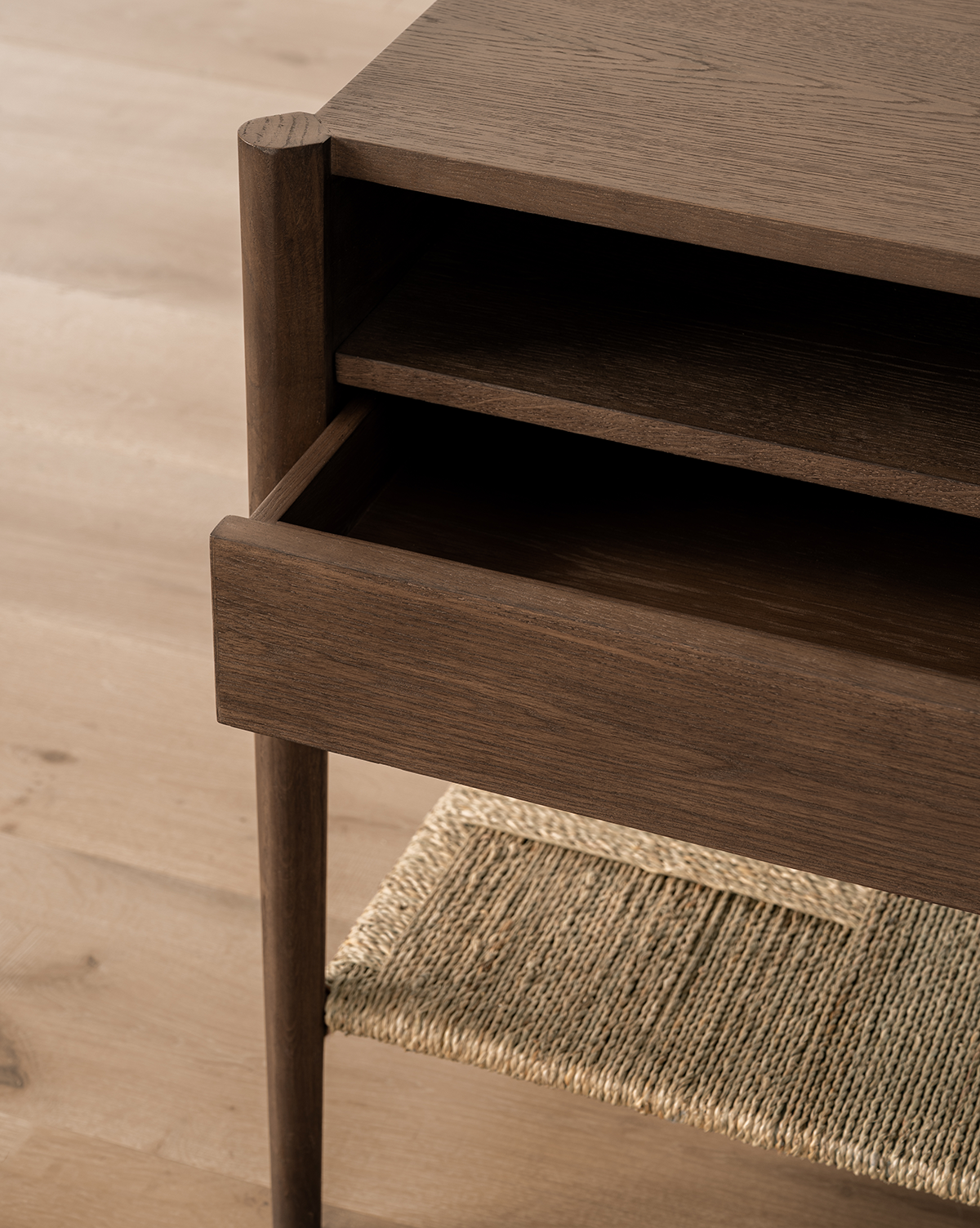 A close-up of the Dana Walnut Oak Nightstand shows its open drawer with brass hardware, shelf above the drawer, and woven rattan lower shelf, all set on light wood flooring, by McGee & Co.