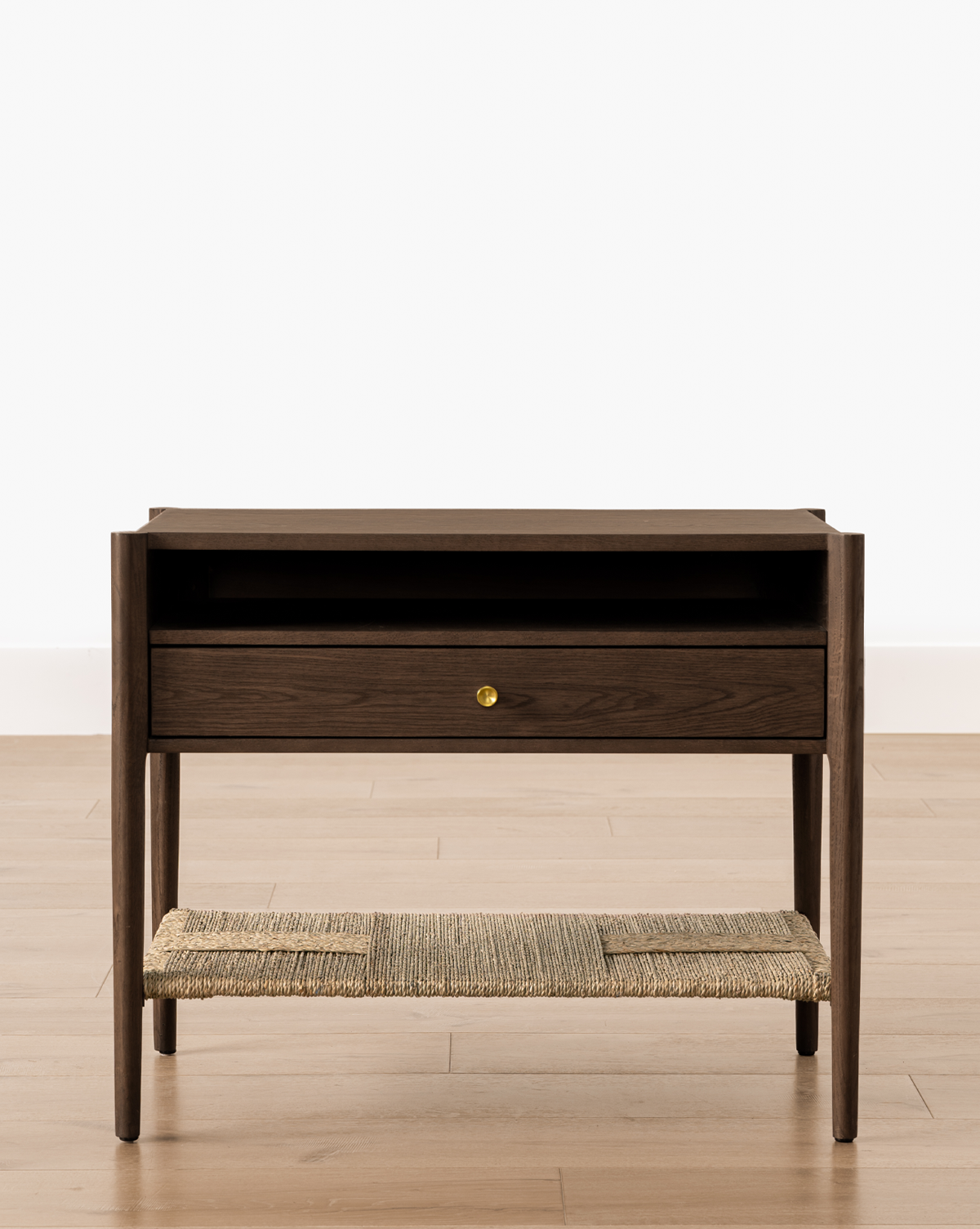 The Dana Walnut Oak Nightstand, featuring brass hardware, a single drawer with a round brass knob, an open shelf above, and a woven rattan lower shelf, stands on a light wooden floor against a white wall, by McGee & Co.