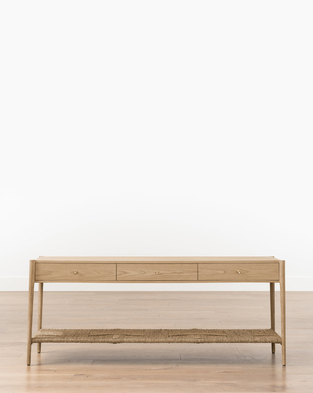 The Dana White Oak Console features a minimalist design with soft close drawers, a woven seagrass shelf, and stands on a wooden floor against a plain white wall, by McGee & Co.