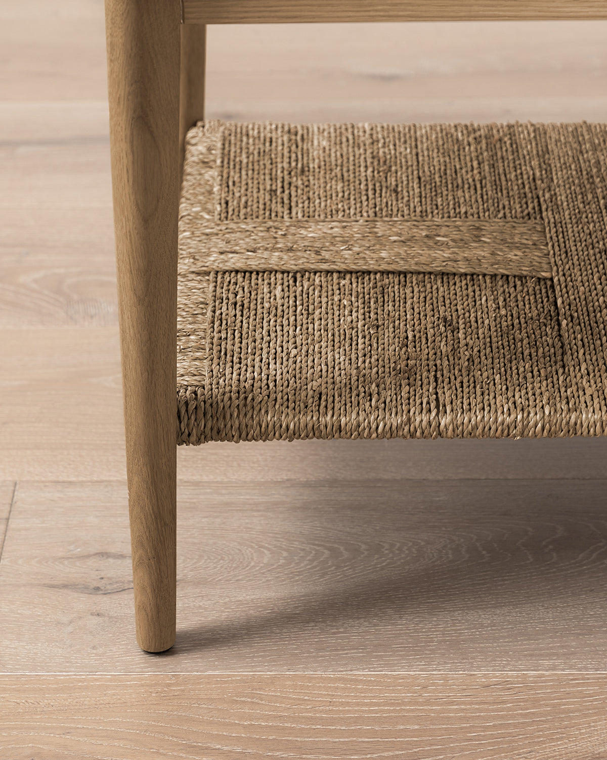 Close-up of a wooden furniture leg beside a woven seagrass shelf on a light wood floor, by McGee & Co.