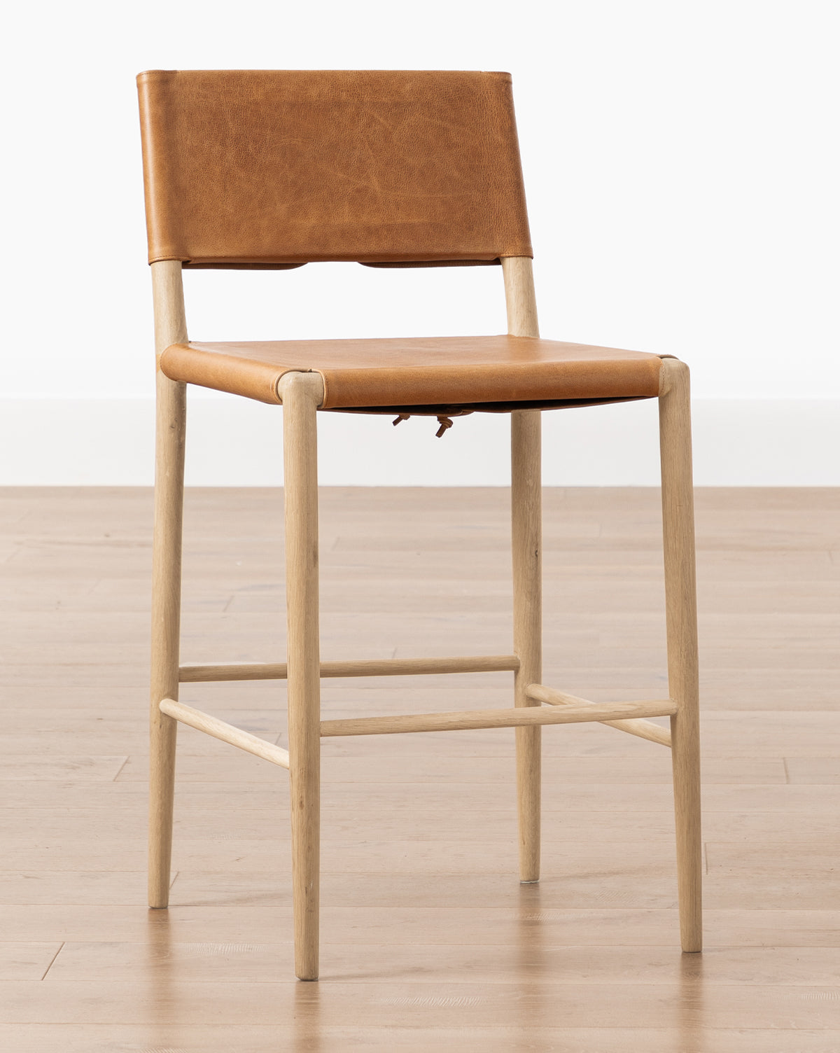The Danson Counter Stool has a minimalist design with a light wooden frame, tan leather seat and backrest, shown on a light wood floor with a plain white background by McGee & Co.