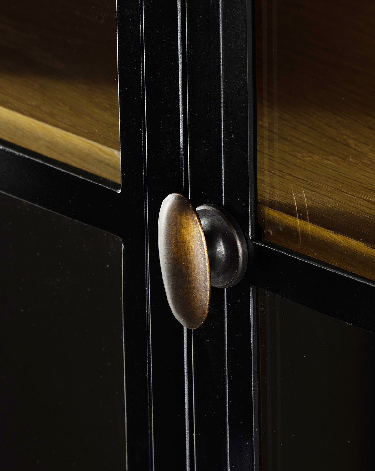 Close-up of the Darcie Cabinet’s antique brass-finished metallic handle, mounted on a black-framed glass door. The glass reflects warm golden-brown tones, enhancing elegance in your home decor.