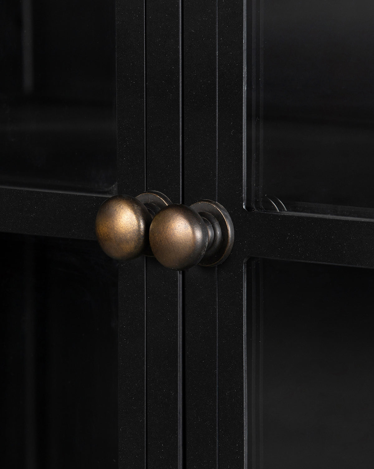 Close-up of two round, antique brass knobs on the Deano Cabinet’s black framed glass doors, showcasing their metallic finish and the elegant intersecting lines of this black hutch, by McGee & Co.