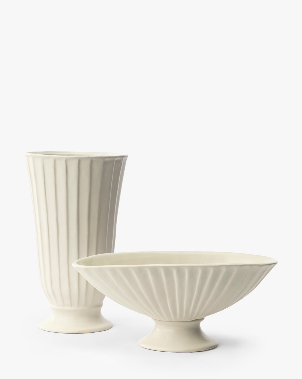 An off-white ceramic set with vertical ridges: a tall, narrow vase stands on the left, and the Delphi Footed Bowl—a wide, fluted vessel with a pedestal base—is placed to the right, both against a plain white background by McGee & Co.