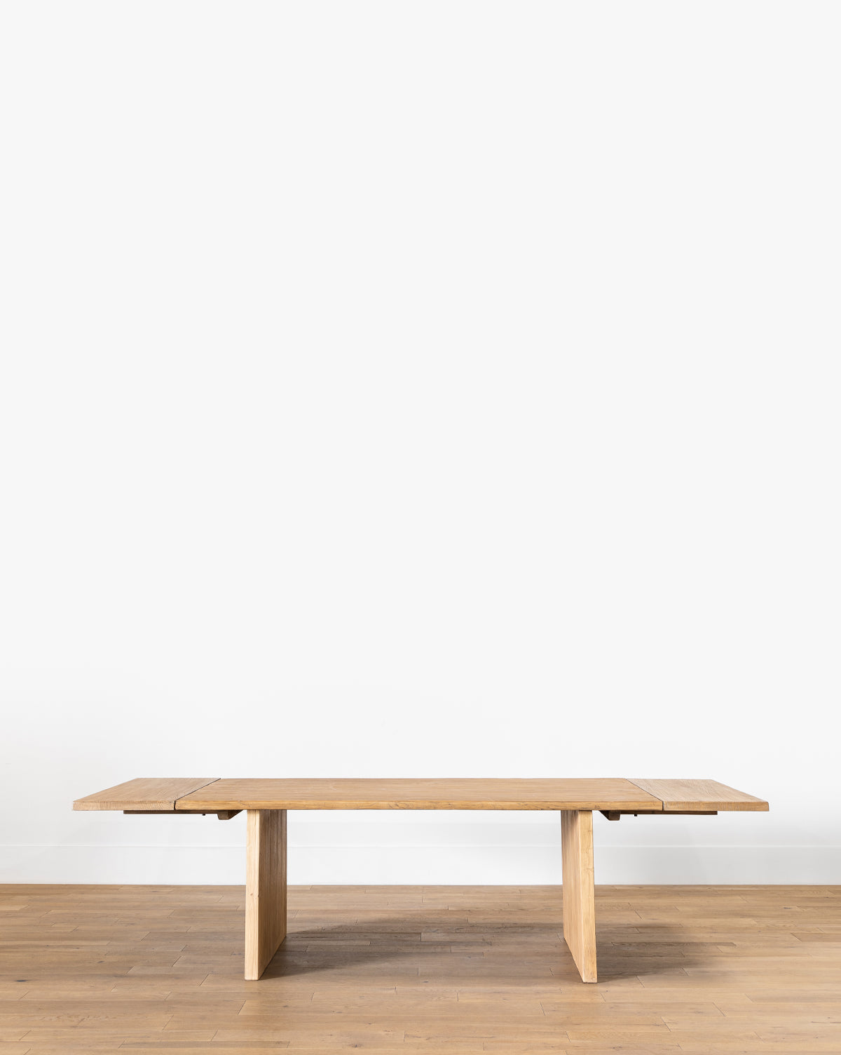 The Dillon Extension Dining Table, a long rectangular piece with simple, clean lines crafted from reclaimed elm, sits on a light wood floor against a plain white wall by McGee & Co.