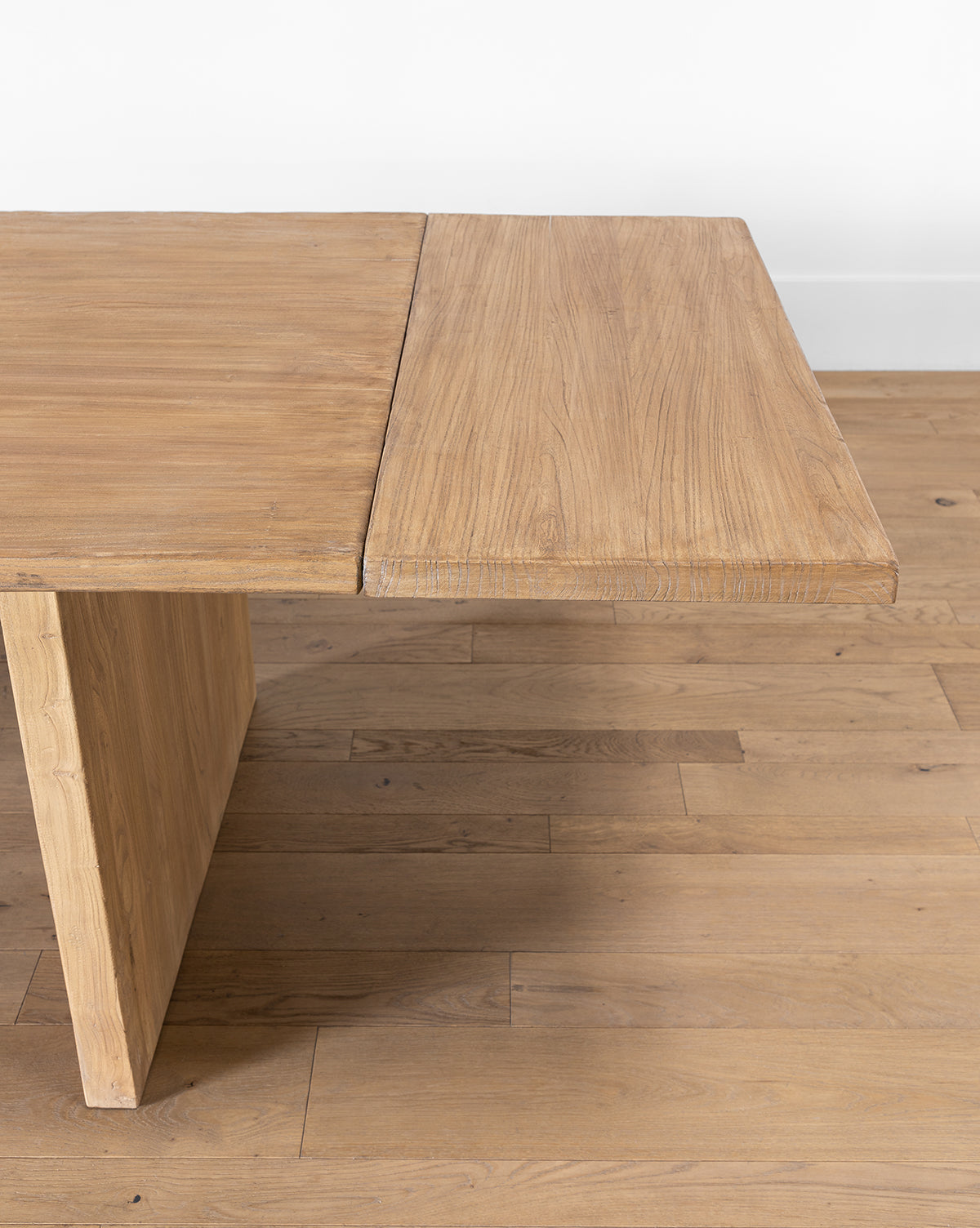 A close-up of the Dillon Extension Dining Table shows its rectangular shape, thick textured top, and angled leg, set on a wooden floor against a white wall by McGee & Co.