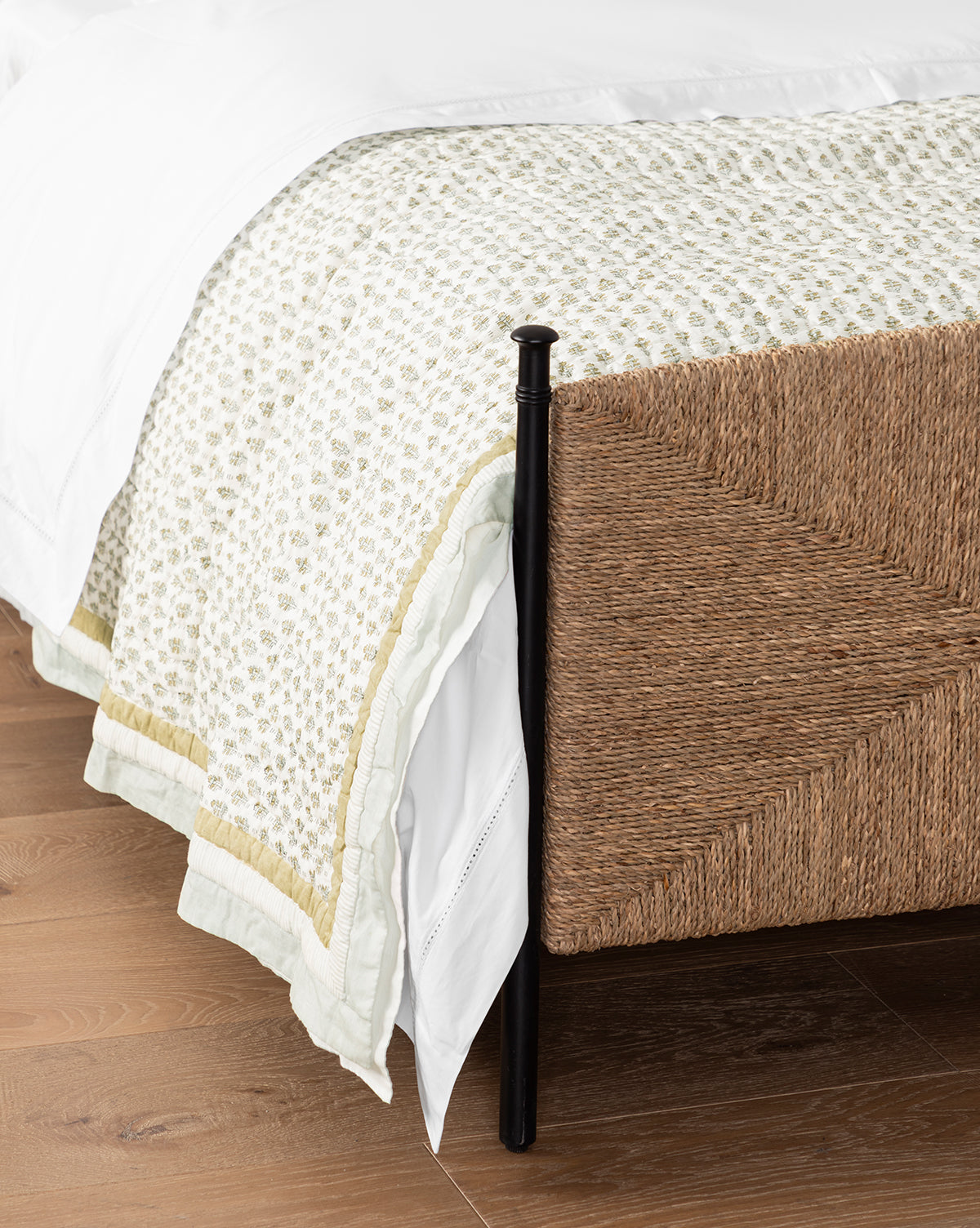 A close-up view of a bed with a woven rattan footboard, white bedding, and the Dulcie Block Print Kids Quilt, set on a wooden floor, by McGee & Co.