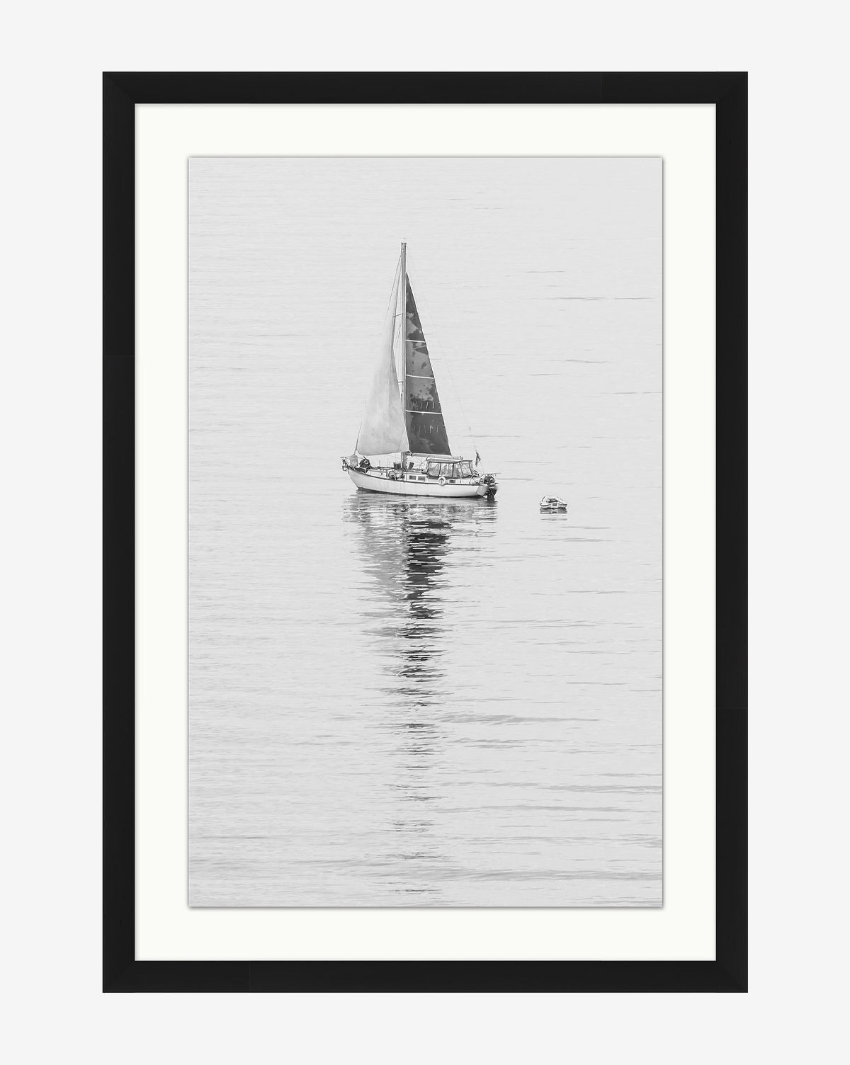 Early Voyage is a black and white photo featuring a sailboat with a single sail gliding on calm water, its reflection visible, with a small dinghy nearby. The striking image is elegantly framed in a simple black wood frame, by McGee & Co.