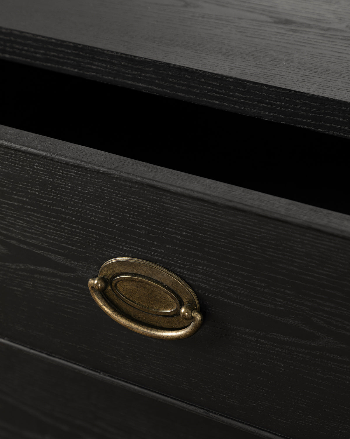 A close-up of the Edward Nightstand shows its Black Ash finish and oval brass handle on a partially open drawer, revealing a dark, empty interior that highlights its modern bedside table design, by McGee & Co.