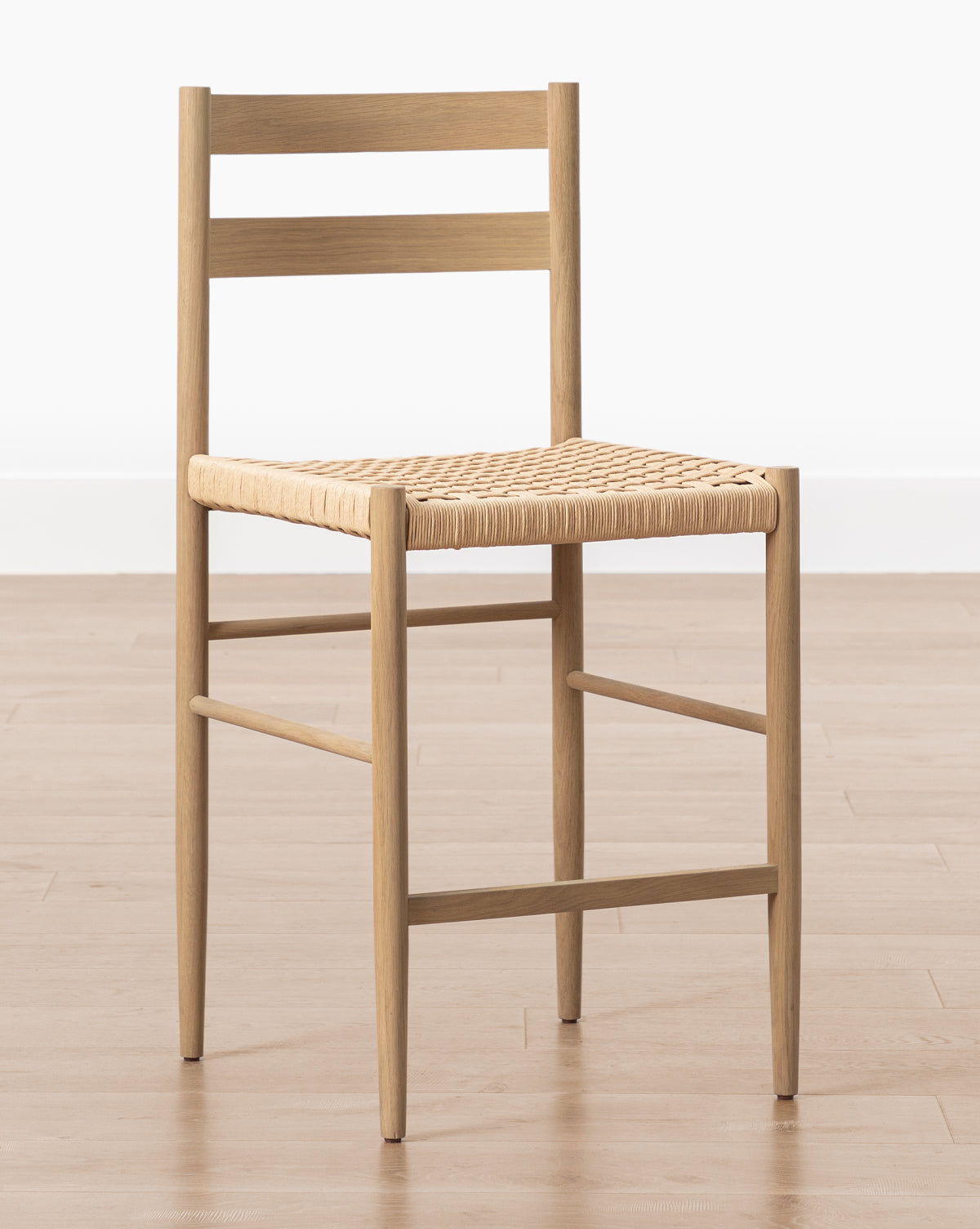 The Eloise Woven Counter Stool features a light oak frame, straight legs, horizontal backrest slats, and a woven seat in classic Danish design, shown on a wooden floor against a plain white wall by McGee & Co.