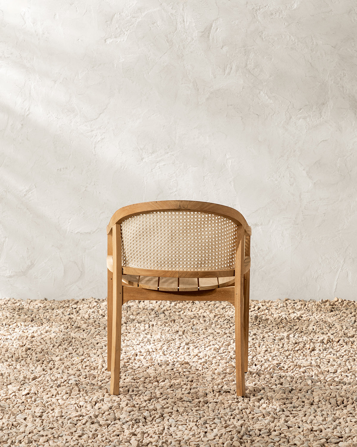 The Elowyn Outdoor Dining Chair, featuring a curved back and woven cane panel, sits on a gravel floor against a textured white wall, illuminated by soft natural light, by McGee & Co.