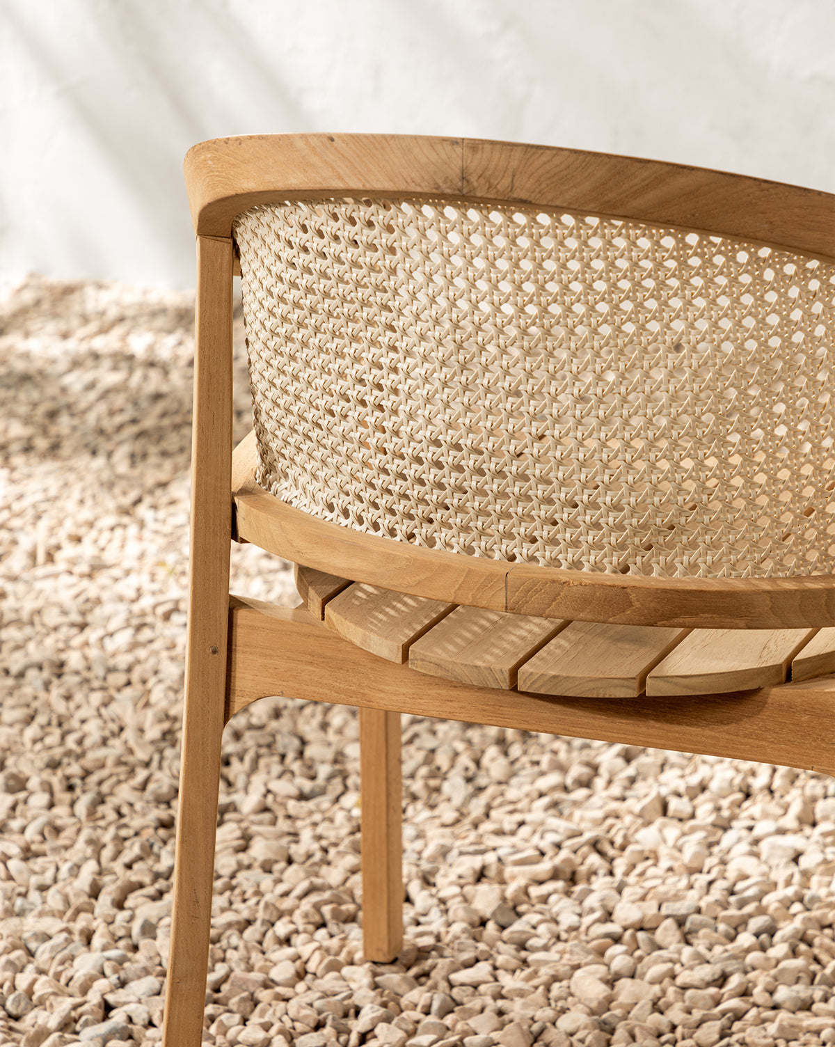 The Elowyn Outdoor Dining Chair, featuring a woven cane backrest and teak frame, is set on light pebbles against a white textured wall illuminated by soft natural light, by McGee & Co.