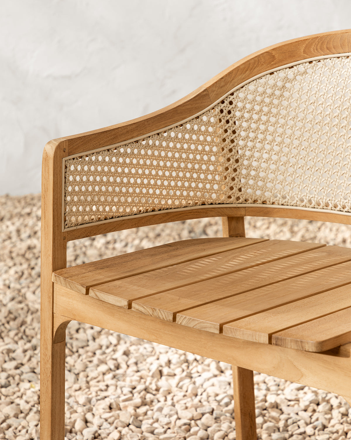 A close-up of the Elowyn Outdoor Dining Chair shows its slatted teak seat and curved backrest with woven cane detailing, set on a surface covered in small, light-colored pebbles, by McGee & Co.