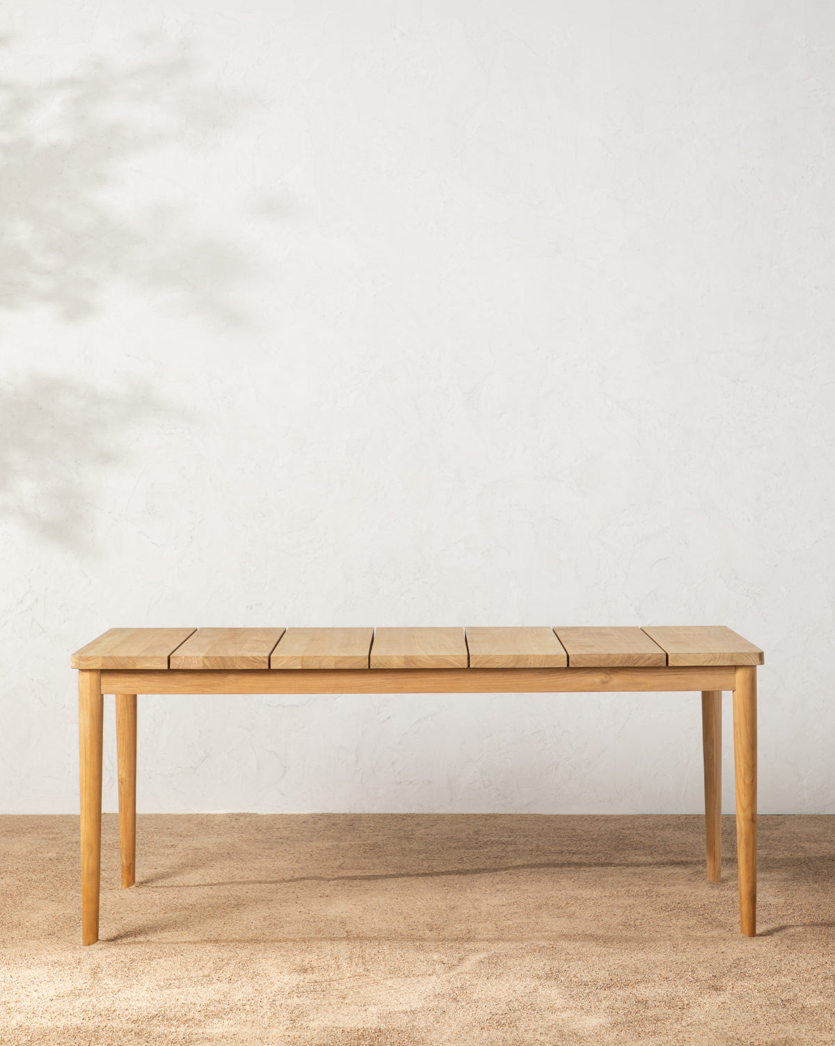 The Elowyn Outdoor Dining Table, made of teak with a slatted top, stands on a textured beige floor against a plain white wall, where soft leaf shadows are cast, by McGee & Co.