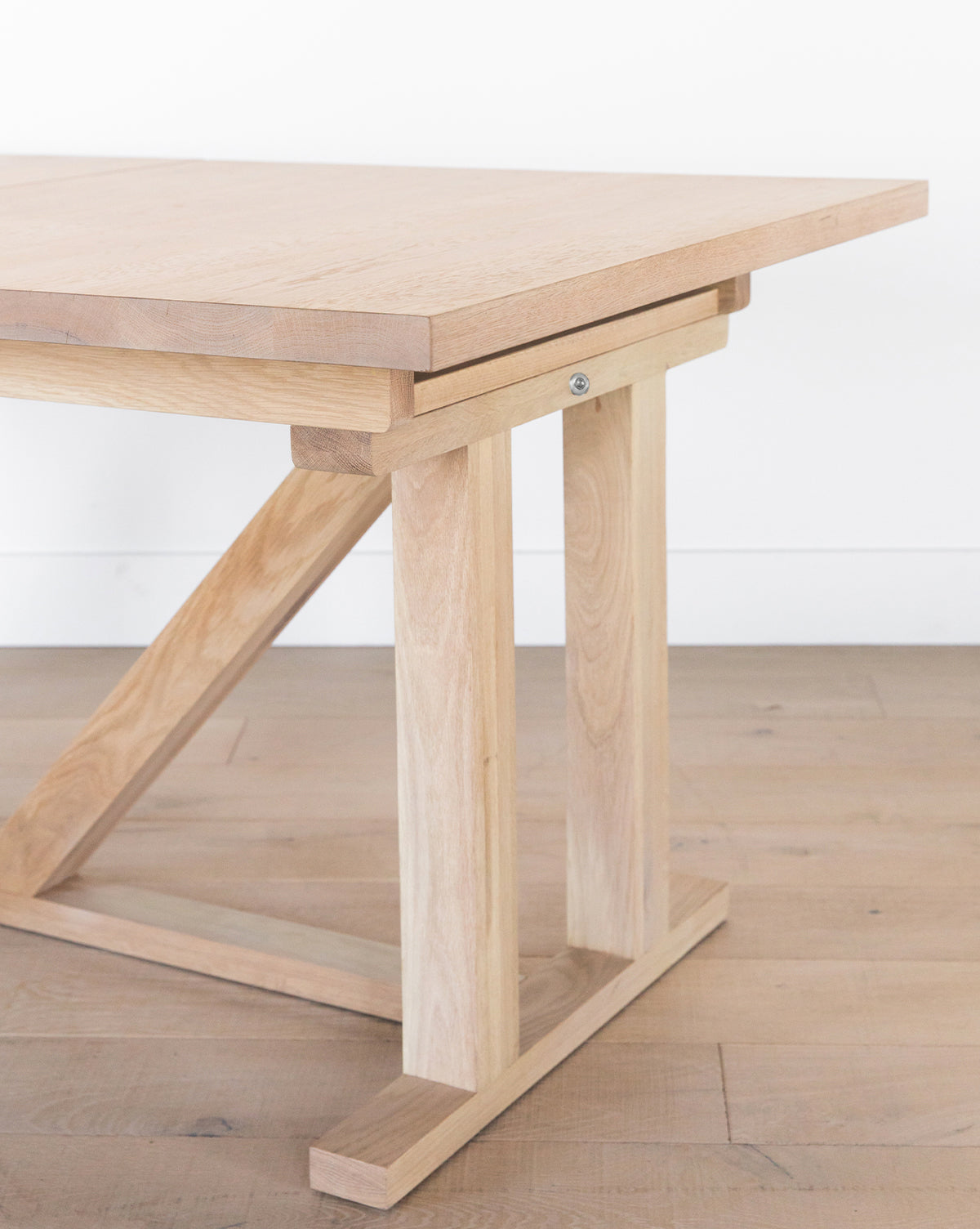 The Emory Extension Dining Table features a light solid oak rectangular top and sturdy, angled architectural base with clean lines, placed on a light wooden floor against a plain white wall by McGee & Co.