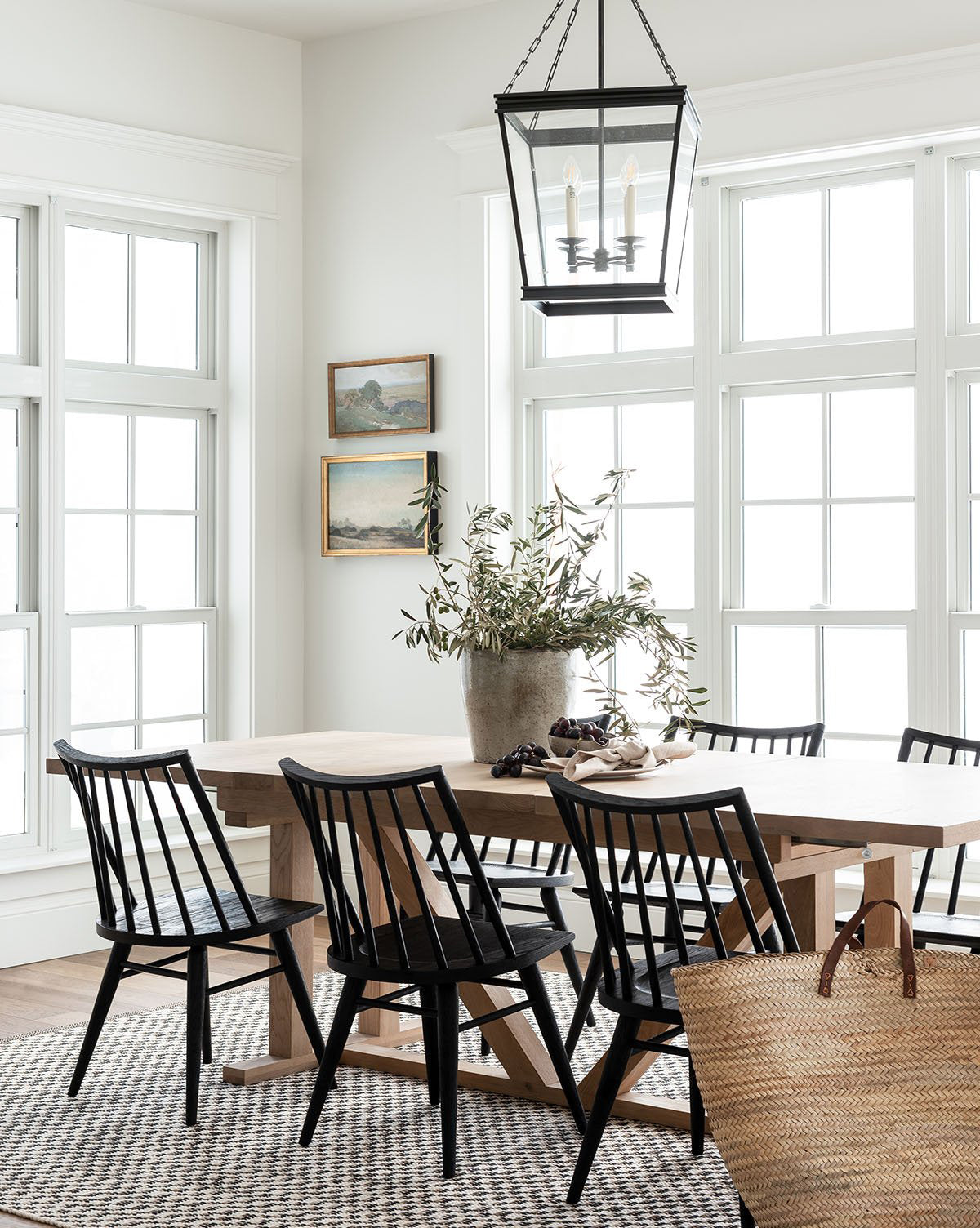 Bright dining area with large windows, an Emory Extension Dining Table, black chairs, potted plant centerpiece, woven rug, two landscape paintings on the wall, and a lantern-style pendant above the table, by McGee & Co.