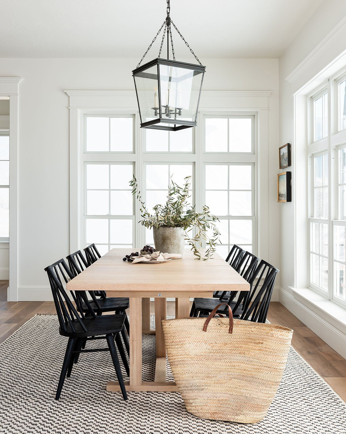 Bright dining room featuring the Emory Extension Dining Table with black chairs, a large potted plant centerpiece, woven basket, patterned rug, and big windows for natural light. A lantern-style light hangs above the table by McGee & Co.