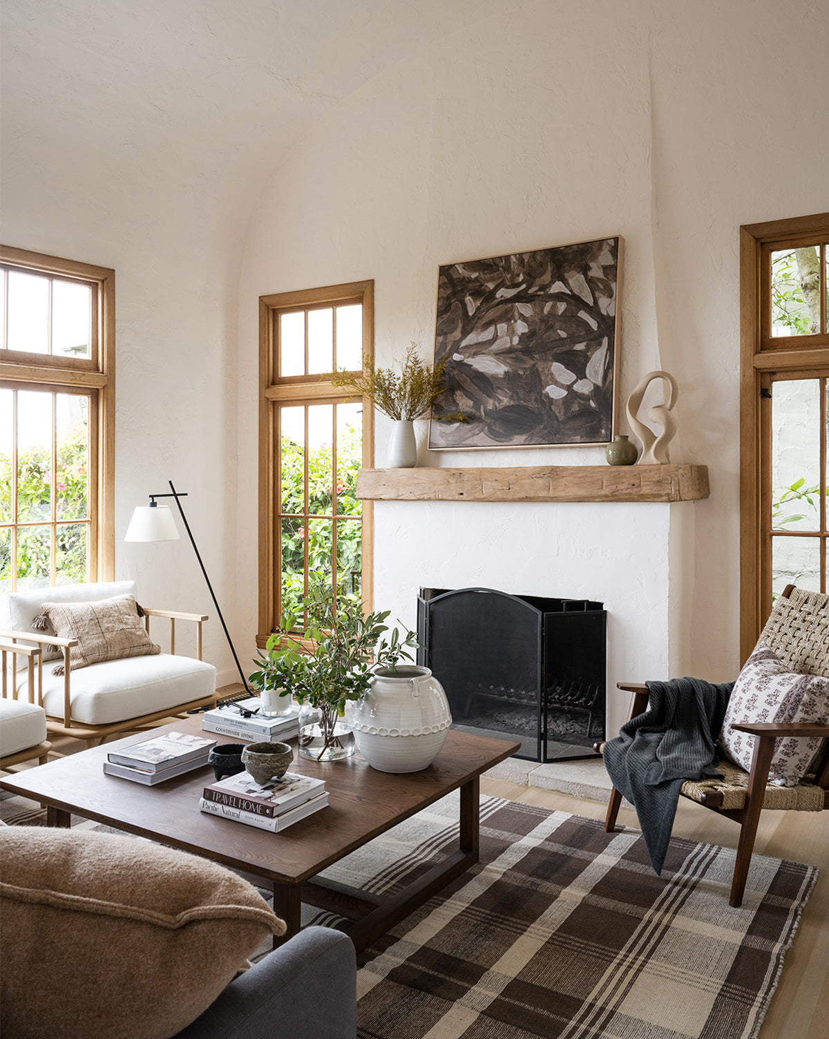 A cozy living room with light walls, wood-framed windows, a Herrin Coffee Table as the centerpiece, neutral armchairs, a plaid rug, and a fireplace with a natural wood mantel. Sunlight fills the space, highlighting tasteful artwork and vases by McGee & Co.