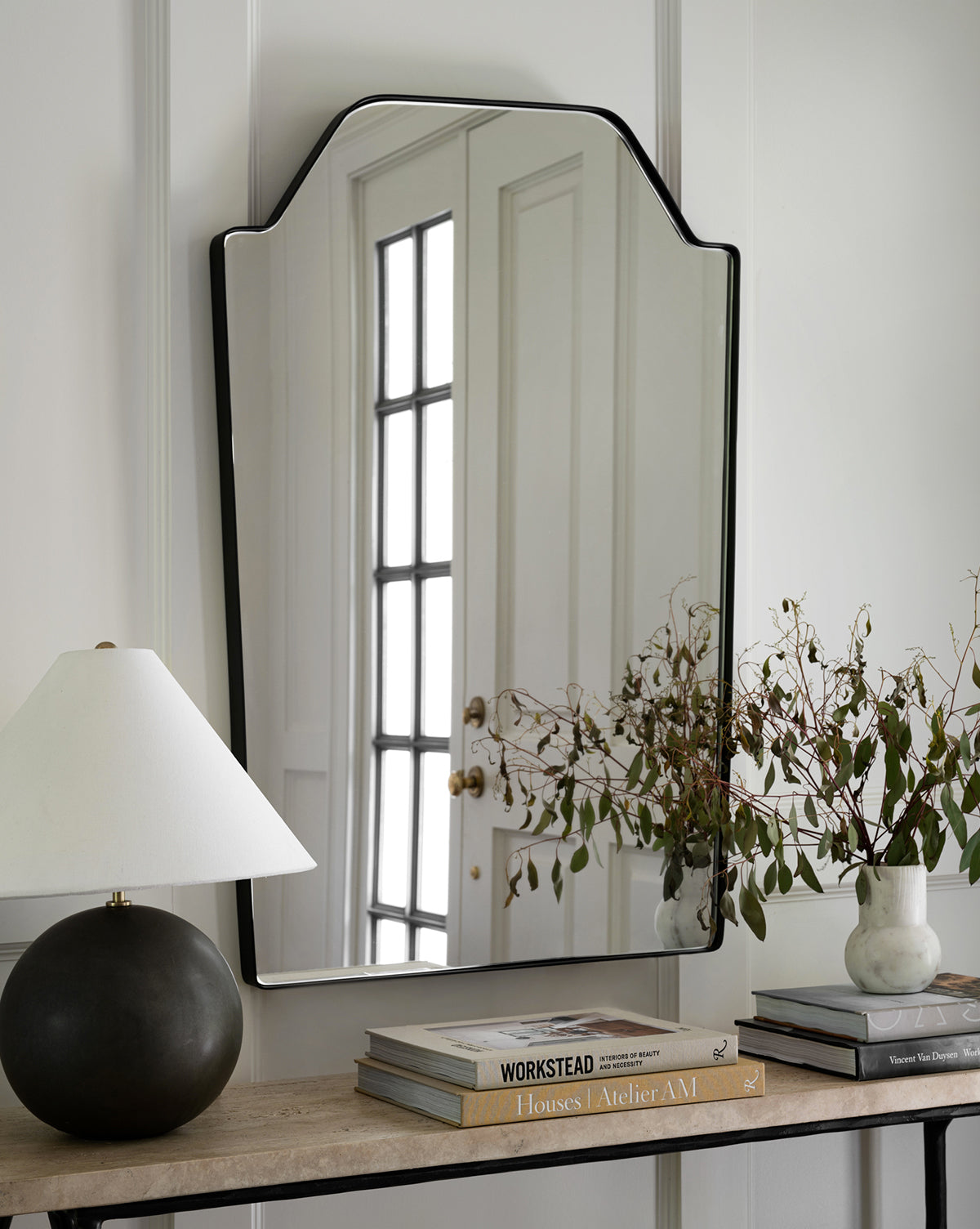 The Giles Wall Mirror with its unique shape hangs above a console table holding a matte black lamp, stacked books, and a vase with leafy branches, while the mirror reflects a door with glass panels by McGee & Co.