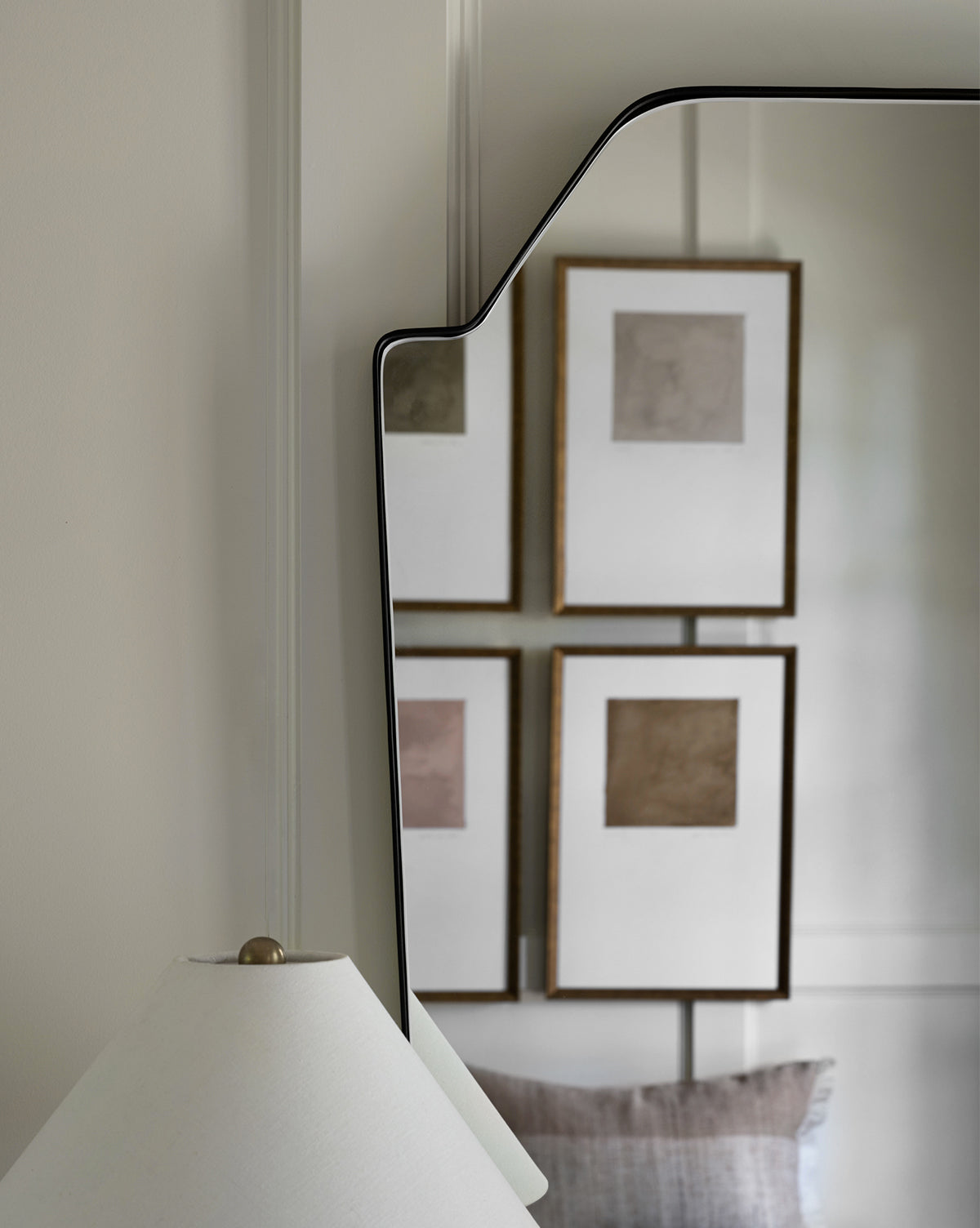 The Giles Wall Mirror reflects four framed abstract artworks, a white lamp with a gold finial, and part of a gray pillow, all set against neutral-toned walls and décor, by McGee & Co.
