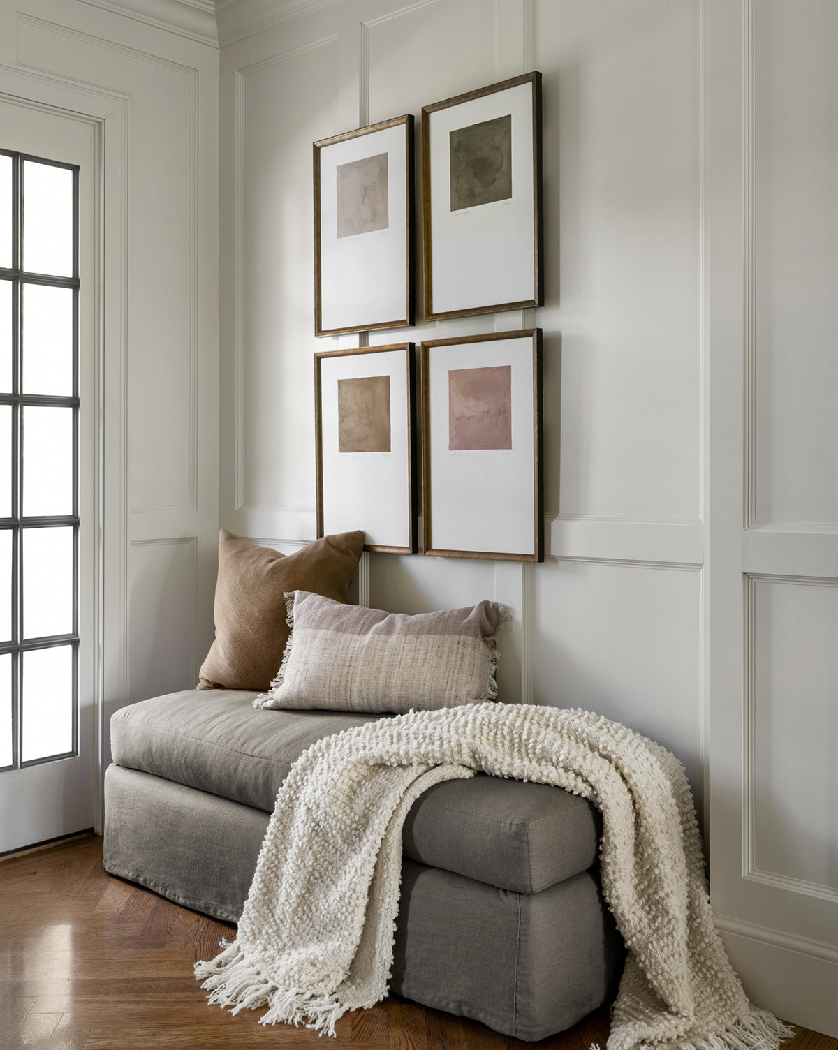 A cozy nook features a cushioned gray bench, neutral throw pillows, a textured cream blanket, and Olive Study minimalist wall art prints above white paneled walls. Natural light streams in through the glass-panel door.