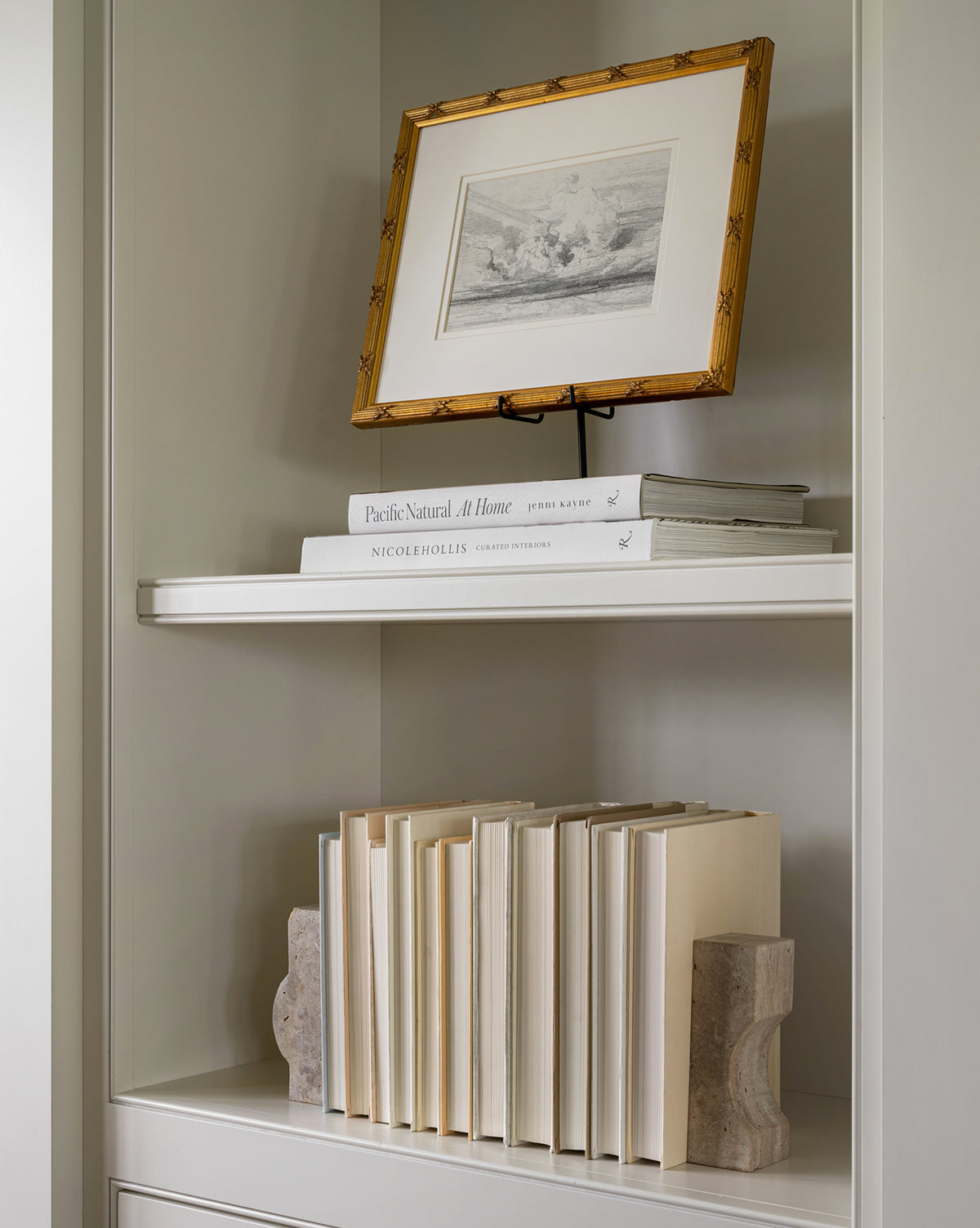 A bookshelf with two shelves: the top displays a framed black-and-white art print and two stacked books, while the lower shelf features cream-colored books upright between decorative Travertine Objects.