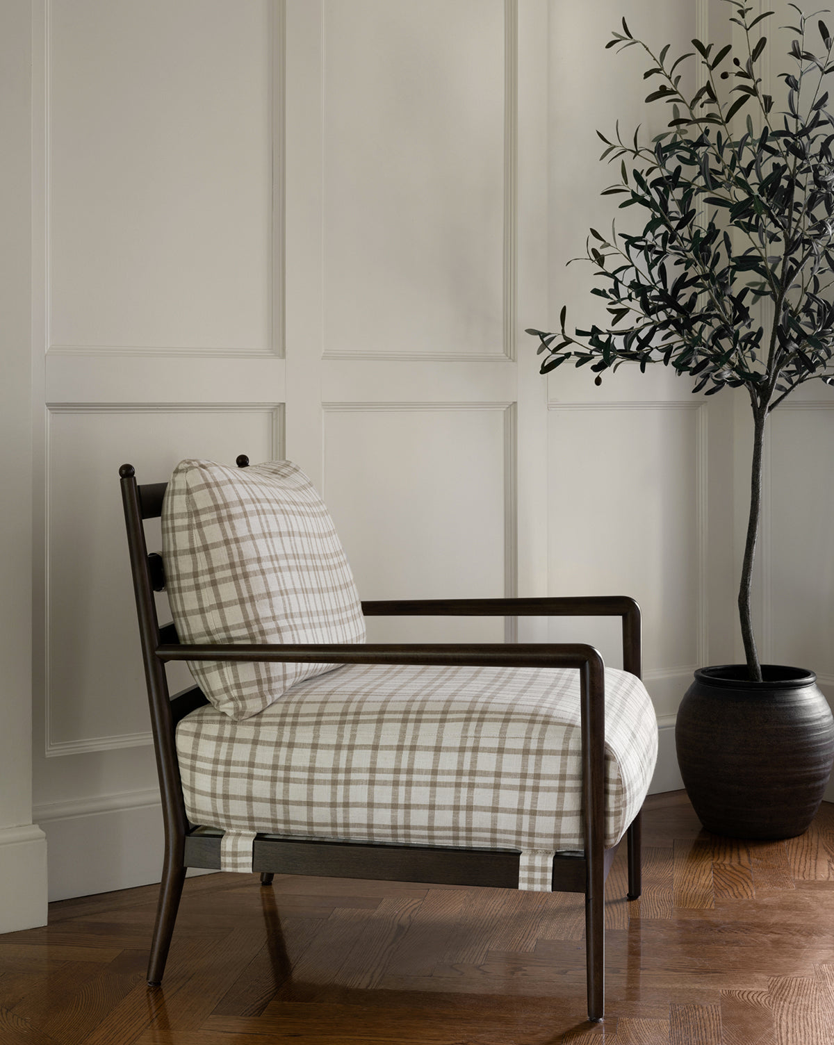 A modern armchair with beige and white plaid upholstery sits on a wooden floor next to the Faux 72 Olive Tree with Ceramic Pot, set against white paneled walls for a minimal and cozy interior look.