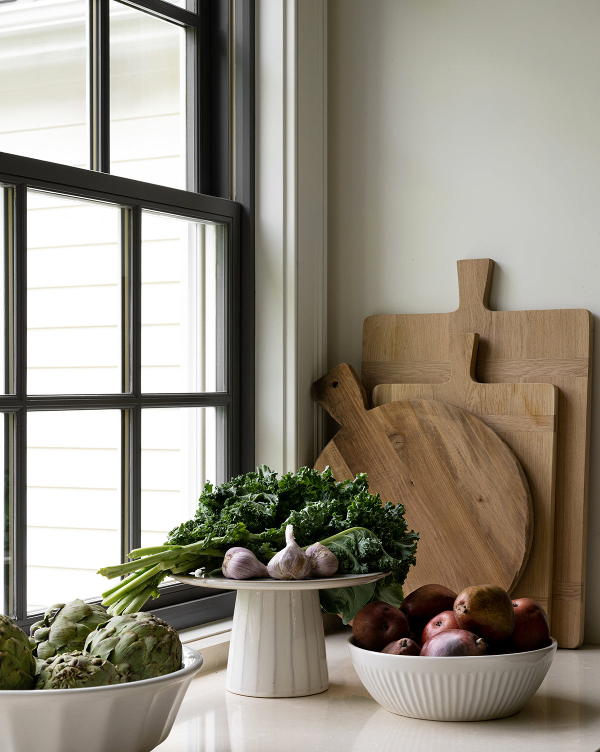 A kitchen countertop by a window displays artichokes in a bowl, leafy greens and garlic on a pedestal, red potatoes in a white bowl, and Rectangle Bread Boards made of oak and walnut with a natural finish propped against the wall, by McGee & Co.