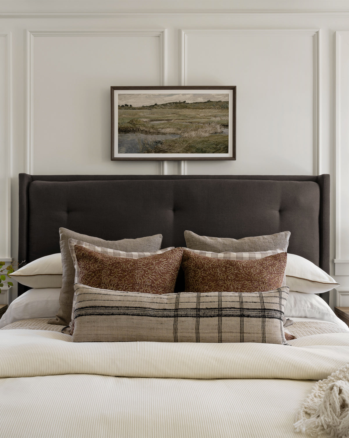 A modern bed with a dark upholstered headboard is set against a white paneled wall, topped with neutral pillows and blankets. Above the bed hangs the Medlock Meadow museum-quality giclee print in a wood frame.