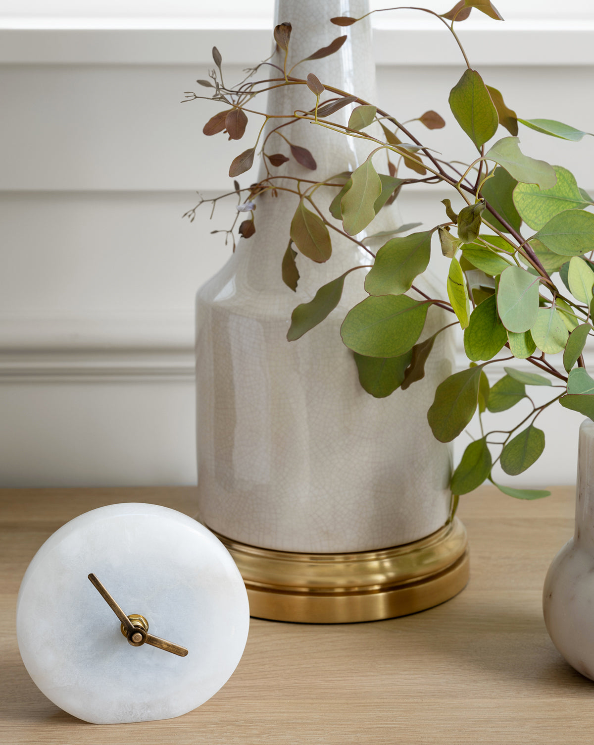 The Alabaster Table Clock with gold hands rests on wood beside a white vase with a gold base and green leaves, offering a sophisticated accent against the white-paneled wall.