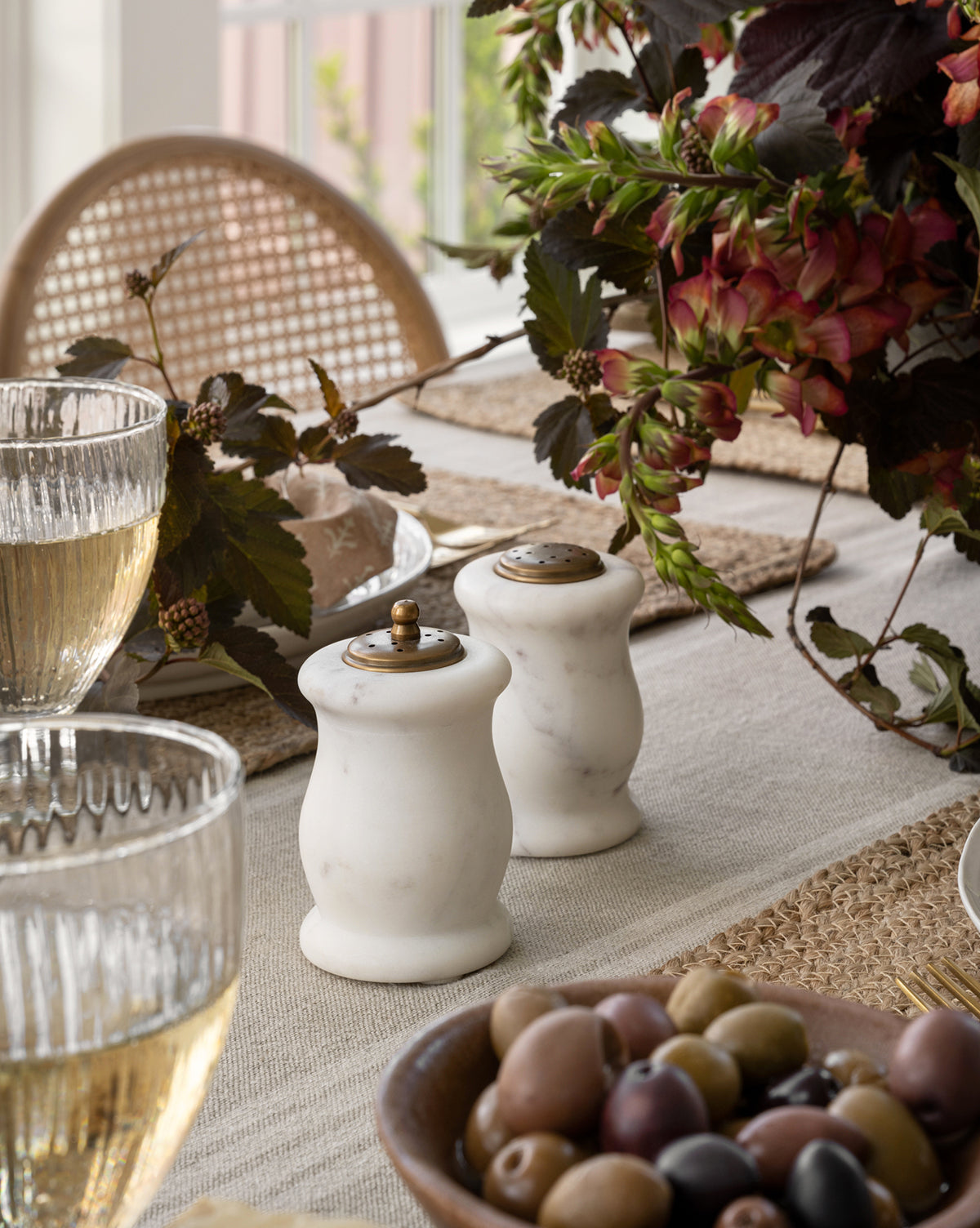 A close-up of a dining table set with glasses of white wine, a bowl of olives, Marble Salt & Pepper Shakers (Set of 2) with antiqued brass toppers, woven placemats, a floral centerpiece, and a rattan chair in the background, by McGee & Co.