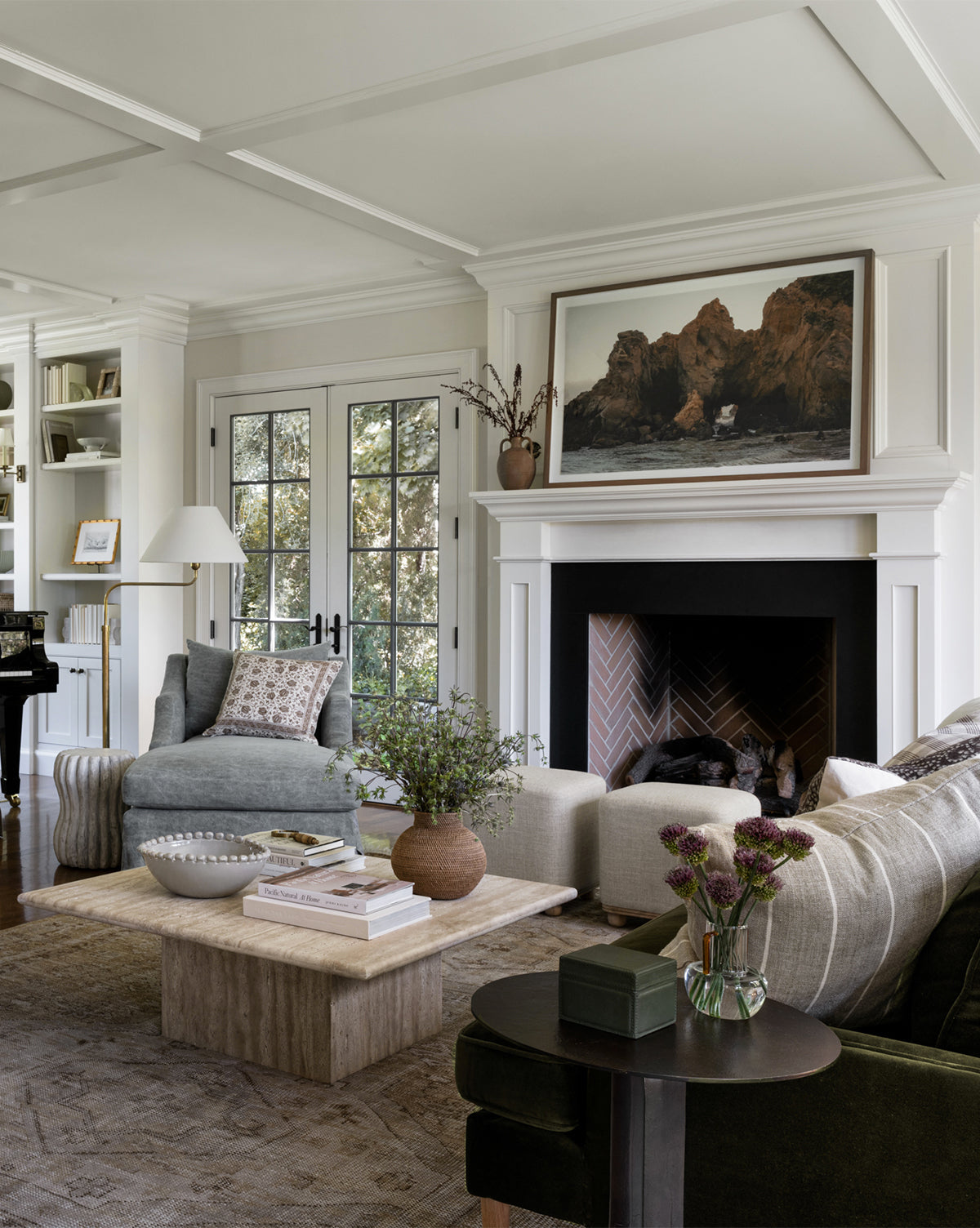 A cozy living room features a fireplace, armchairs, green sofa, and stone coffee table with books and decor. Above the mantel hangs Rock Formation, striking wall art. Large windows brighten the space; shelves line the back wall.