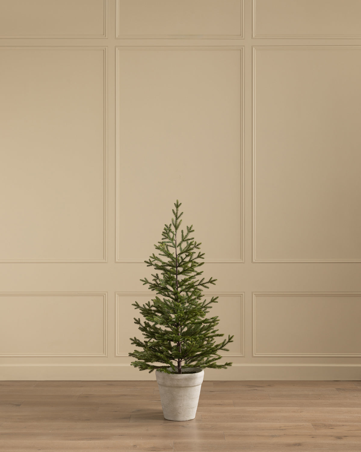 The Faux Spruce Pre-Lit Potted Tree sits on a wooden floor by a beige paneled wall, bringing evergreen charm to your space, by McGee & Co.
