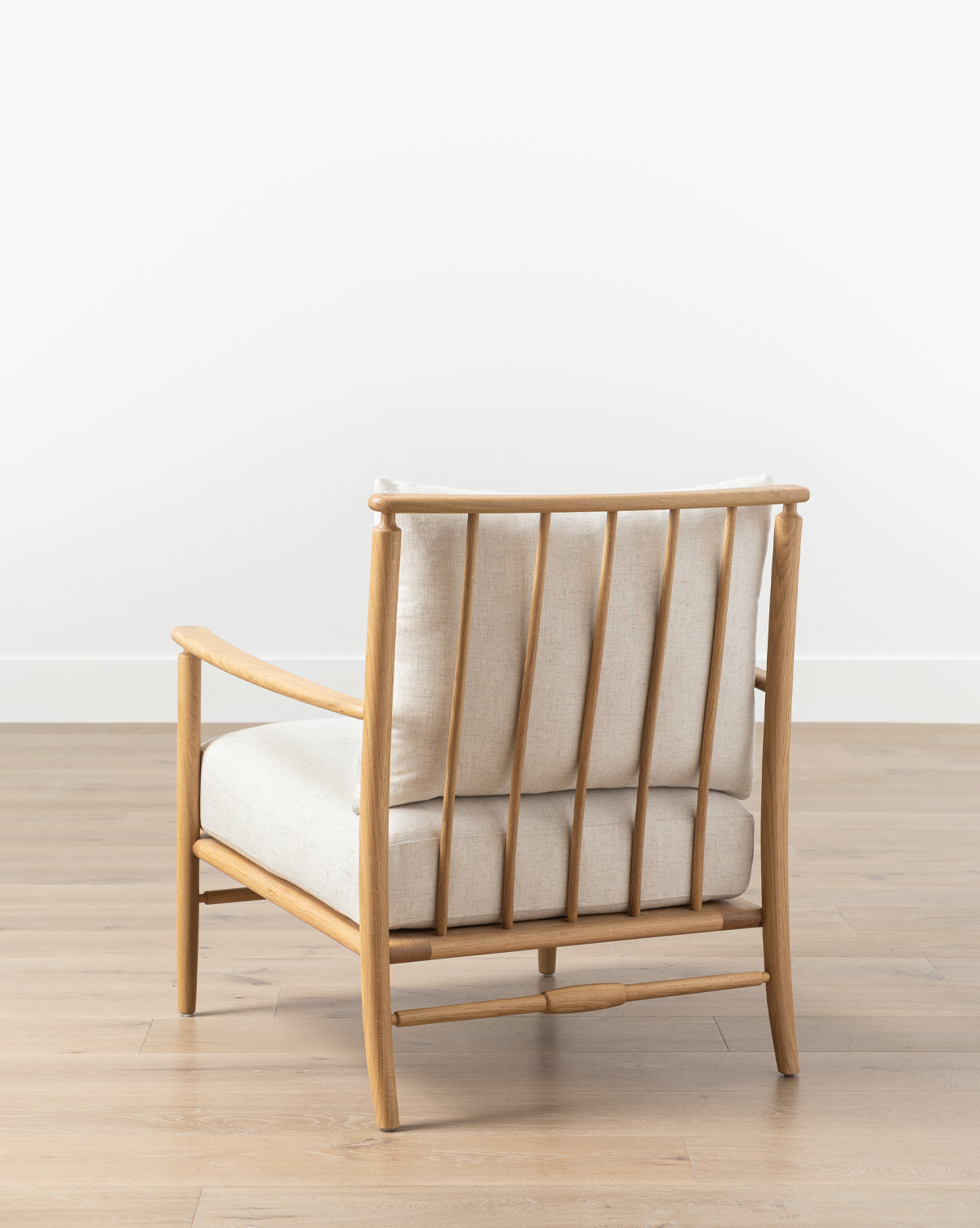 The Fenwick Lounge Chair boasts a modern look with a solid oak frame, vertical back slats, and beige linen cushions—ideal for placement on light wood flooring against a white wall, by McGee & Co.