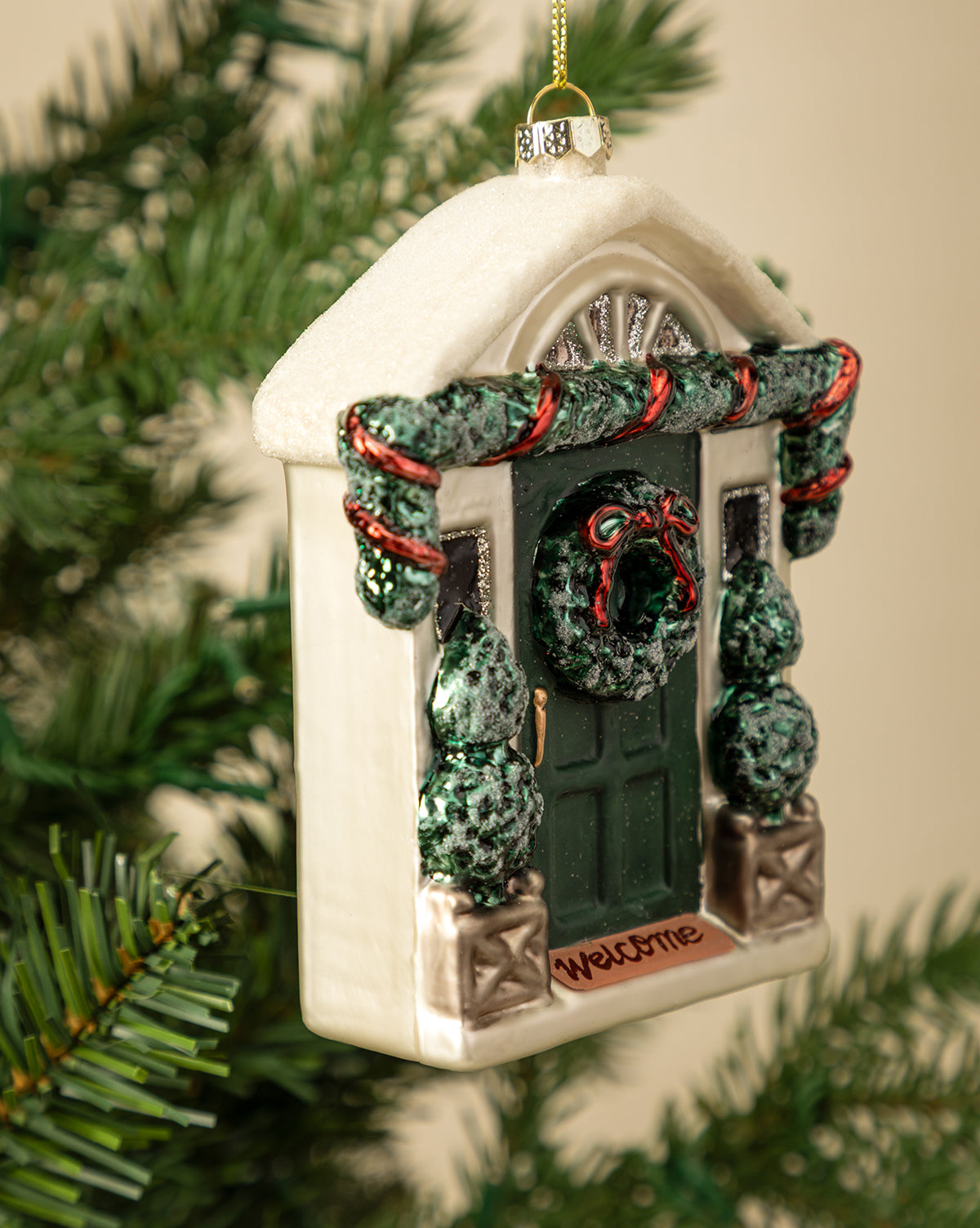 The Festive Front Door Ornament, shaped like a house door with a wreath, garland, and a "Welcome" sign, hangs on a green Christmas tree branch for a charming holiday touch, by McGee & Co.