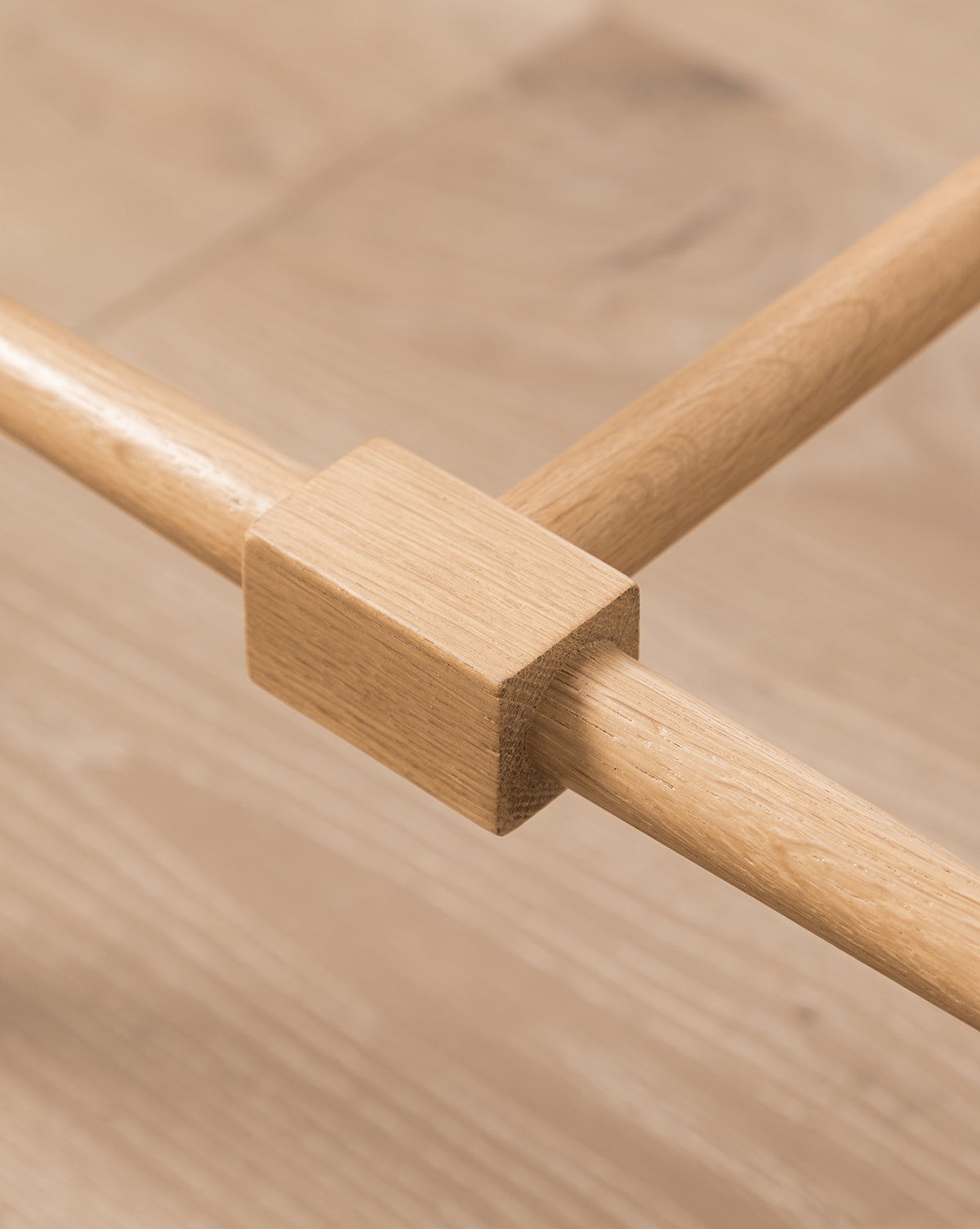 Close-up of a wooden joint on the Fielder Counter Stool, showing a square block joining two cylindrical rods in a light oak finish, set against a light wood floor by McGee & Co.