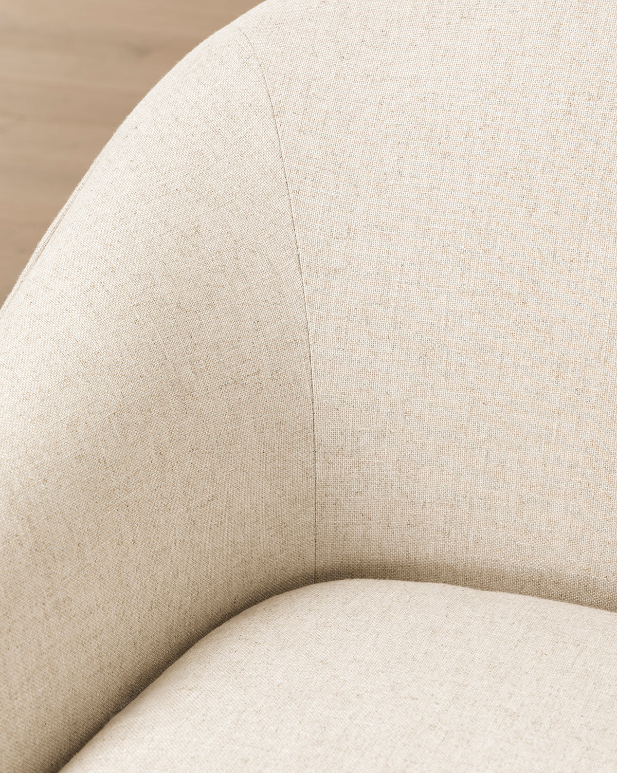 Close-up of the Fletcher Lounge Chair (Ready to Ship) featuring a curved backrest, visible stitching, and smooth textured fabric, by McGee & Co. The customizable beige chair is shown on a wooden floor.