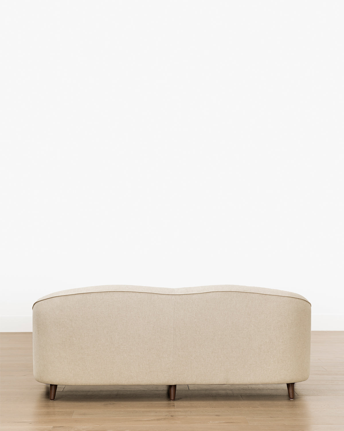 The Fletcher Sofa 76 (Ready to Ship), shown from the back, features a curved back, wooden legs, and beige upholstery on a light wooden floor against a plain white background. Handcrafted and available in customizable fabrics by McGee & Co.