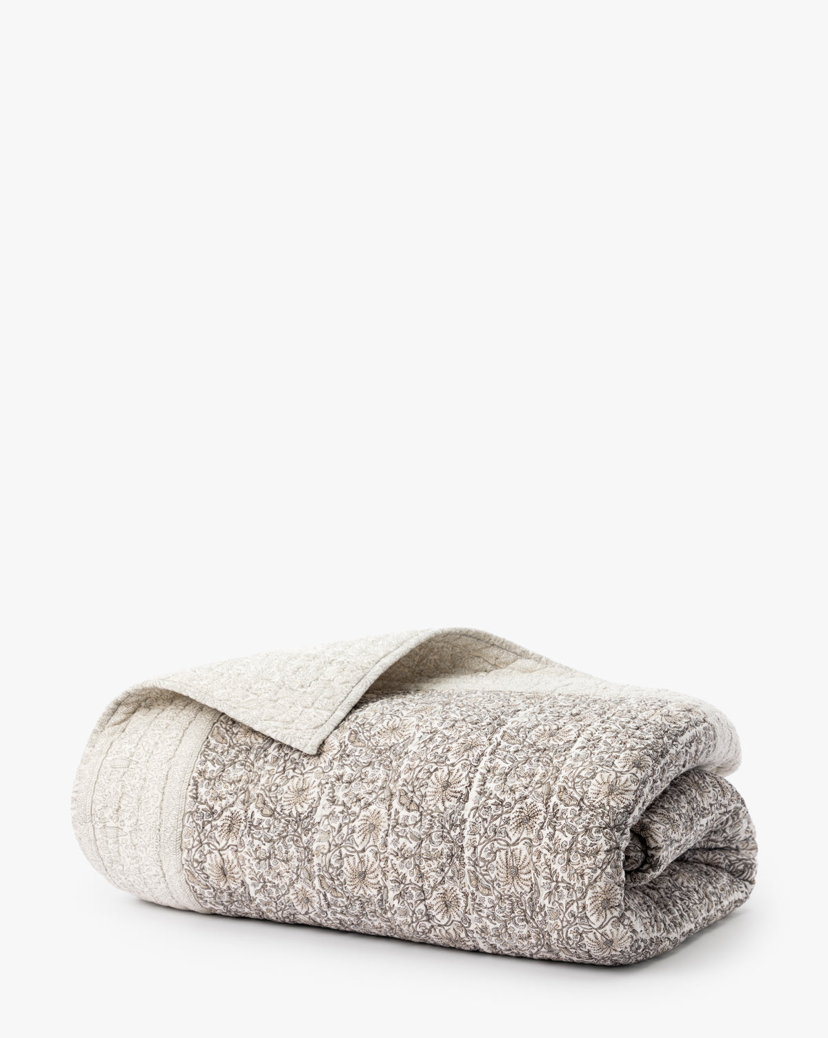 The Floral Block Print Quilt, with a light beige and grey floral pattern, is shown folded and partly open on a white background—an elegant piece of block printed bedding.