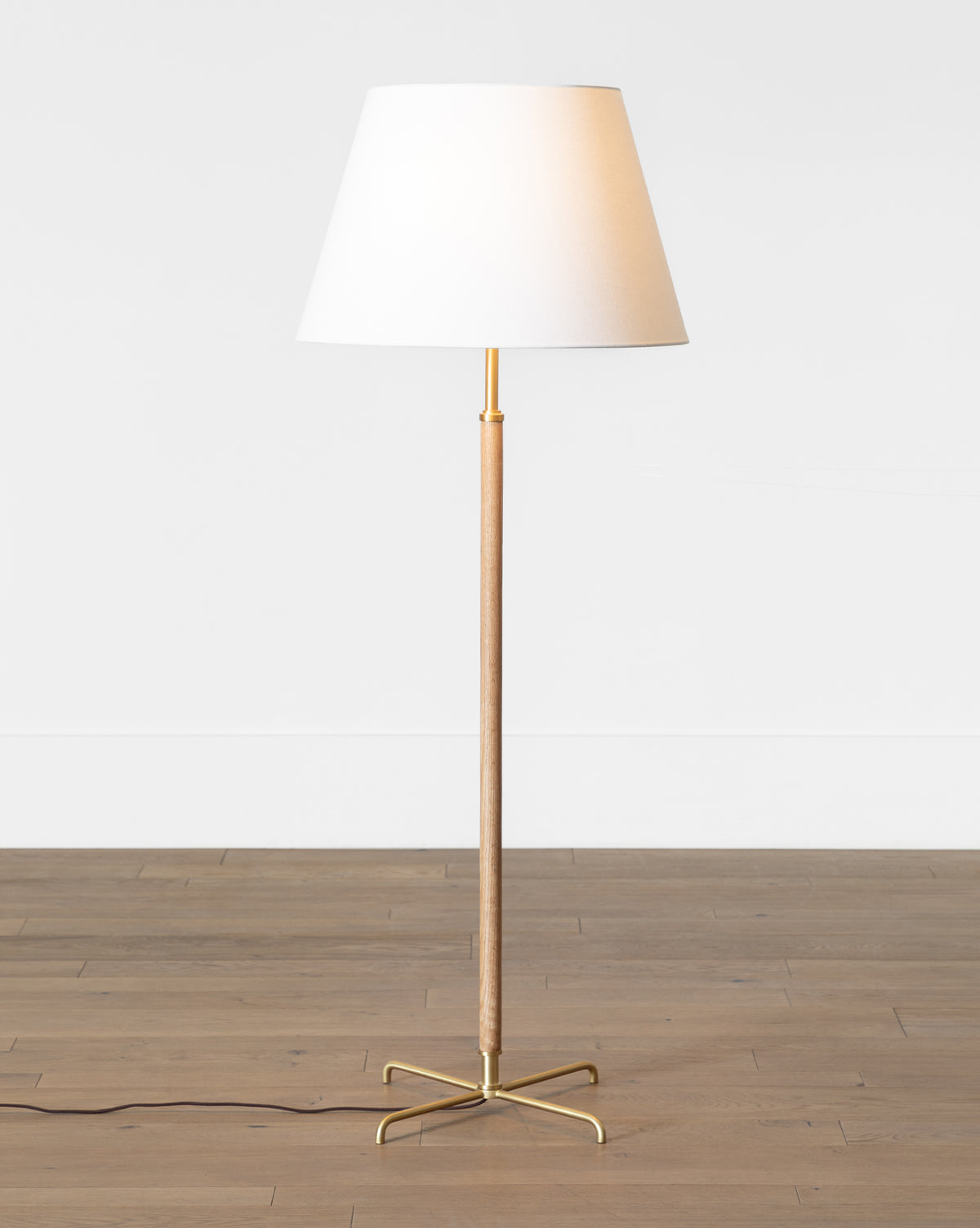The Florence Floor Lamp has a white lampshade, wooden pole, and brass base set on a light wood floor by a plain white wall, with a black power cord extending from the base, by McGee & Co.