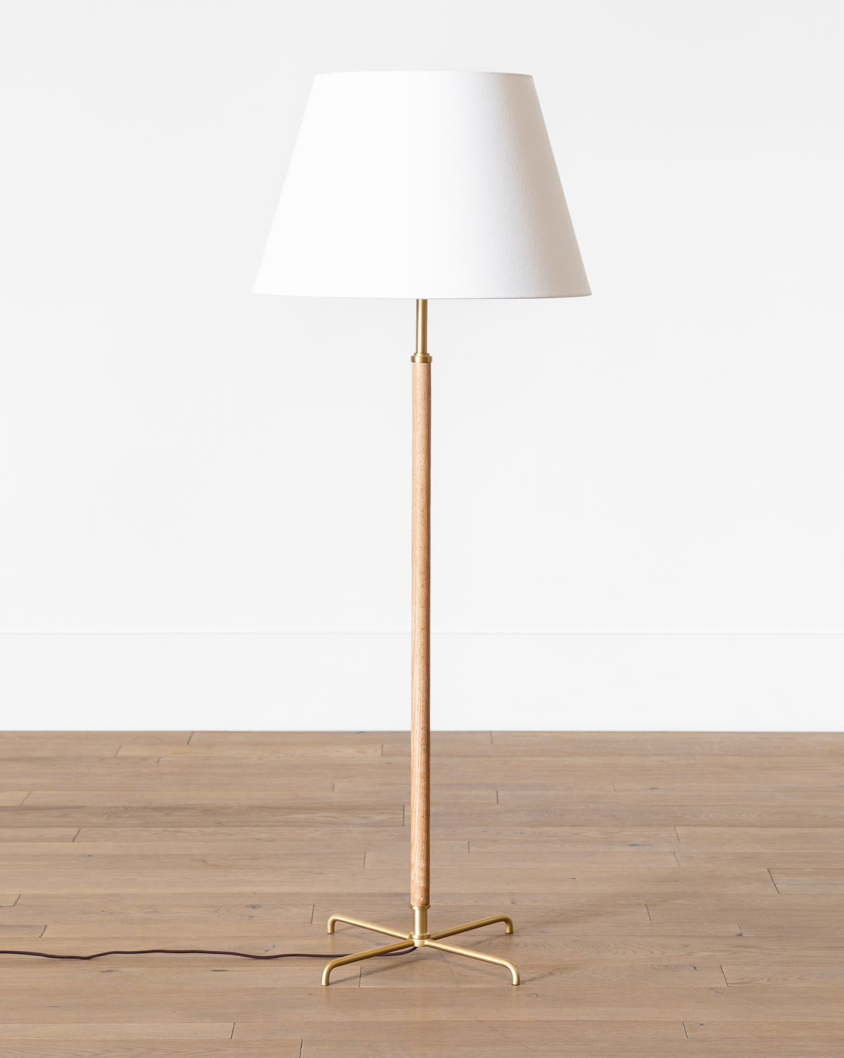 The Makerspalm Florence Floor Lamp features a slim wooden stand, gold metal base, and elegant linen shade, standing on a wooden floor against a plain white wall. McGee & Co.