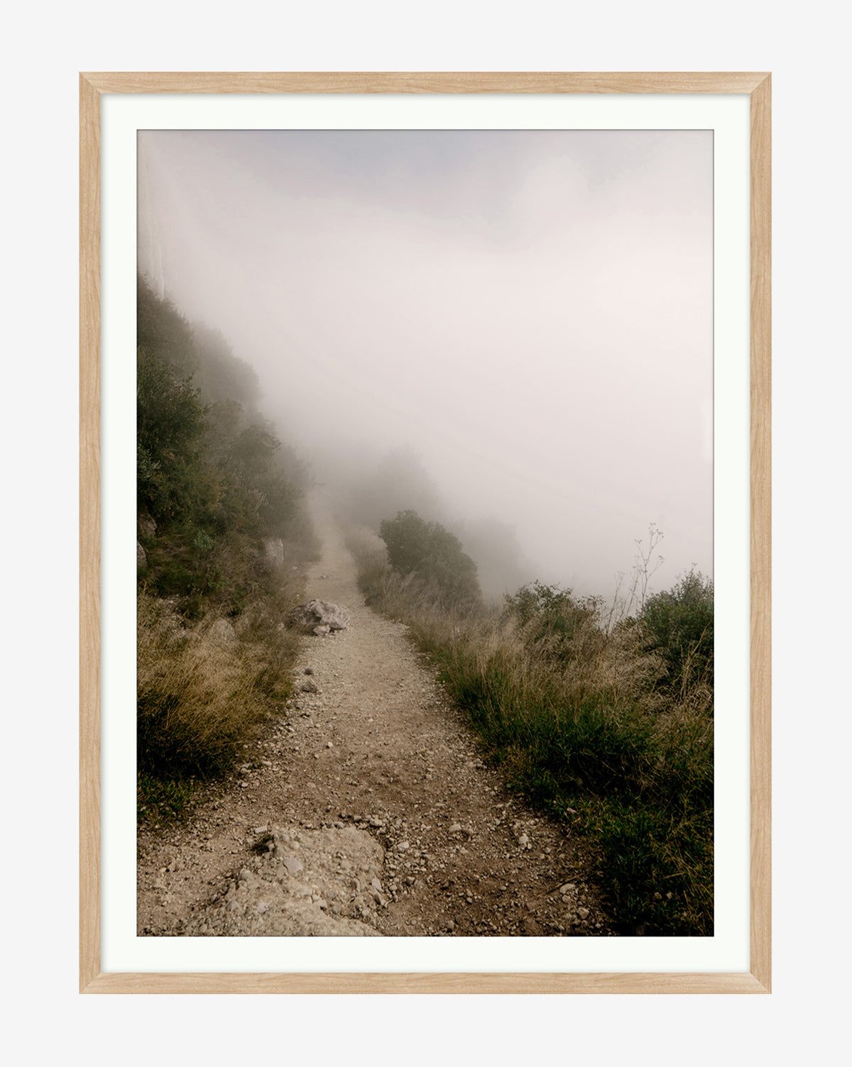 A narrow dirt path winds through grass and rocks, fading into thick white fog as bushes line the misty way—creating a mysterious, tranquil scene reminiscent of the Foggy Path giclee print.