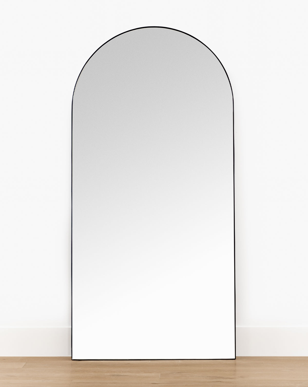 The Gala Floor Mirror features a spacious arched design with a slim black frame, shown leaning against a white wall on wooden flooring in a minimalist setting, by McGee & Co.