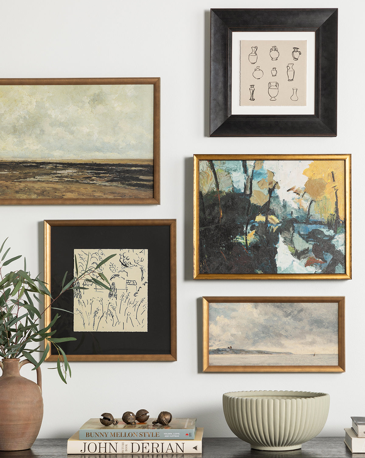 A gallery wall with five framed artworks—including landscapes, the Dark Blue Abstract, and minimalist line drawings in gold wood frames—hangs above a table topped with a potted plant, decorative bowl, and stacked books.
