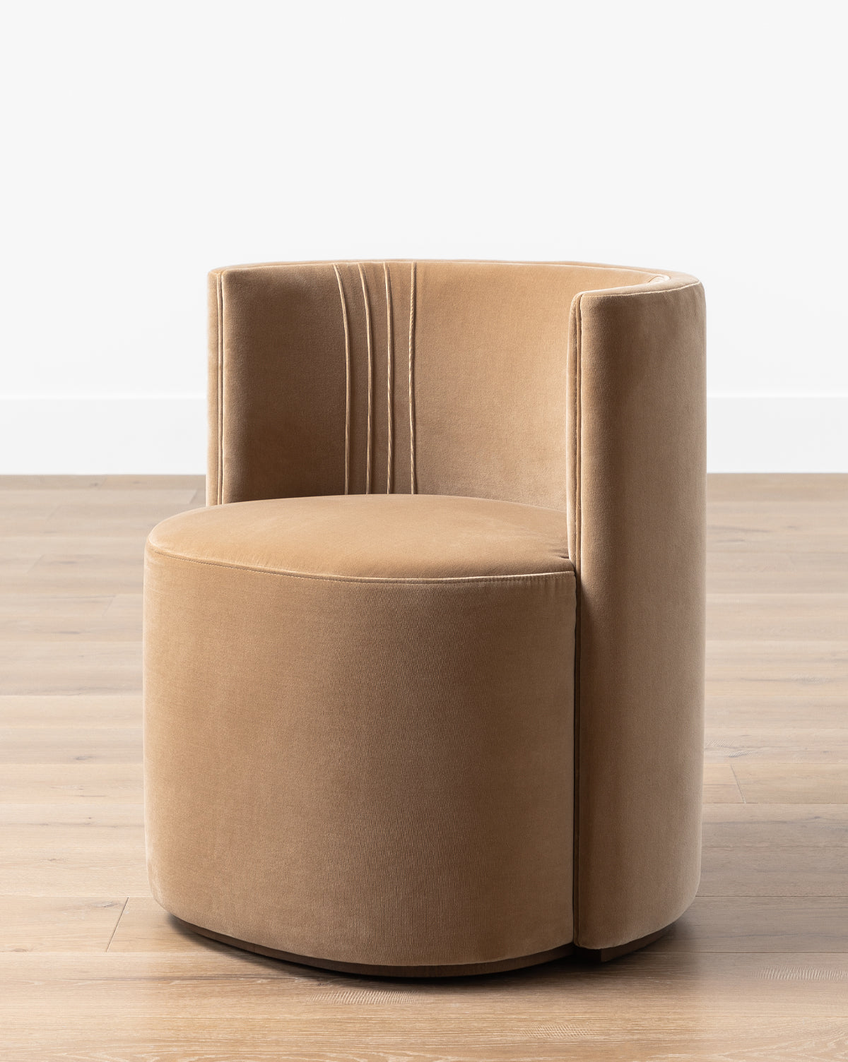 The Georgina Dining Chair (Ready to Ship) features tan velvet upholstery, a barrel back with art deco-inspired vertical stitching, and is shown on a light wooden floor against a plain white wall by McGee & Co.