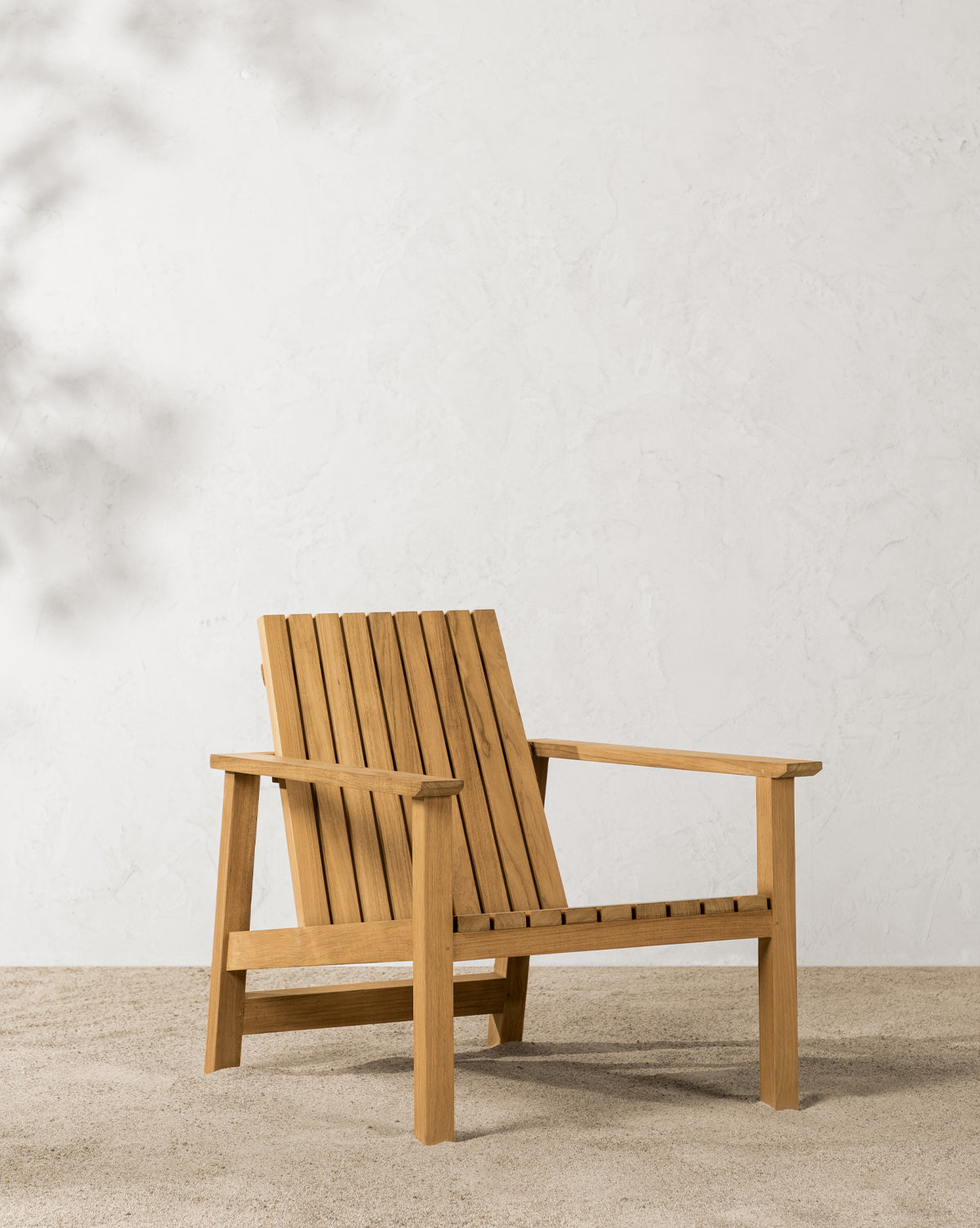 The Gilda Outdoor Lounge Chair, made of teak with a slatted back and seat, is set on gravel against a textured, light-toned wall in natural sunlight by McGee & Co.