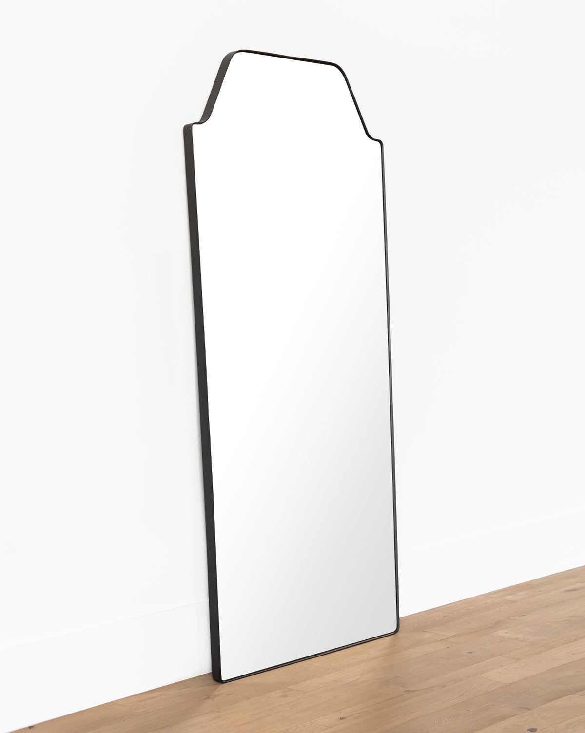 The Giles Floor Mirror, featuring a tall rectangular design with a matte black frame and angled top, leans against a white wall on a wooden floor by McGee & Co.