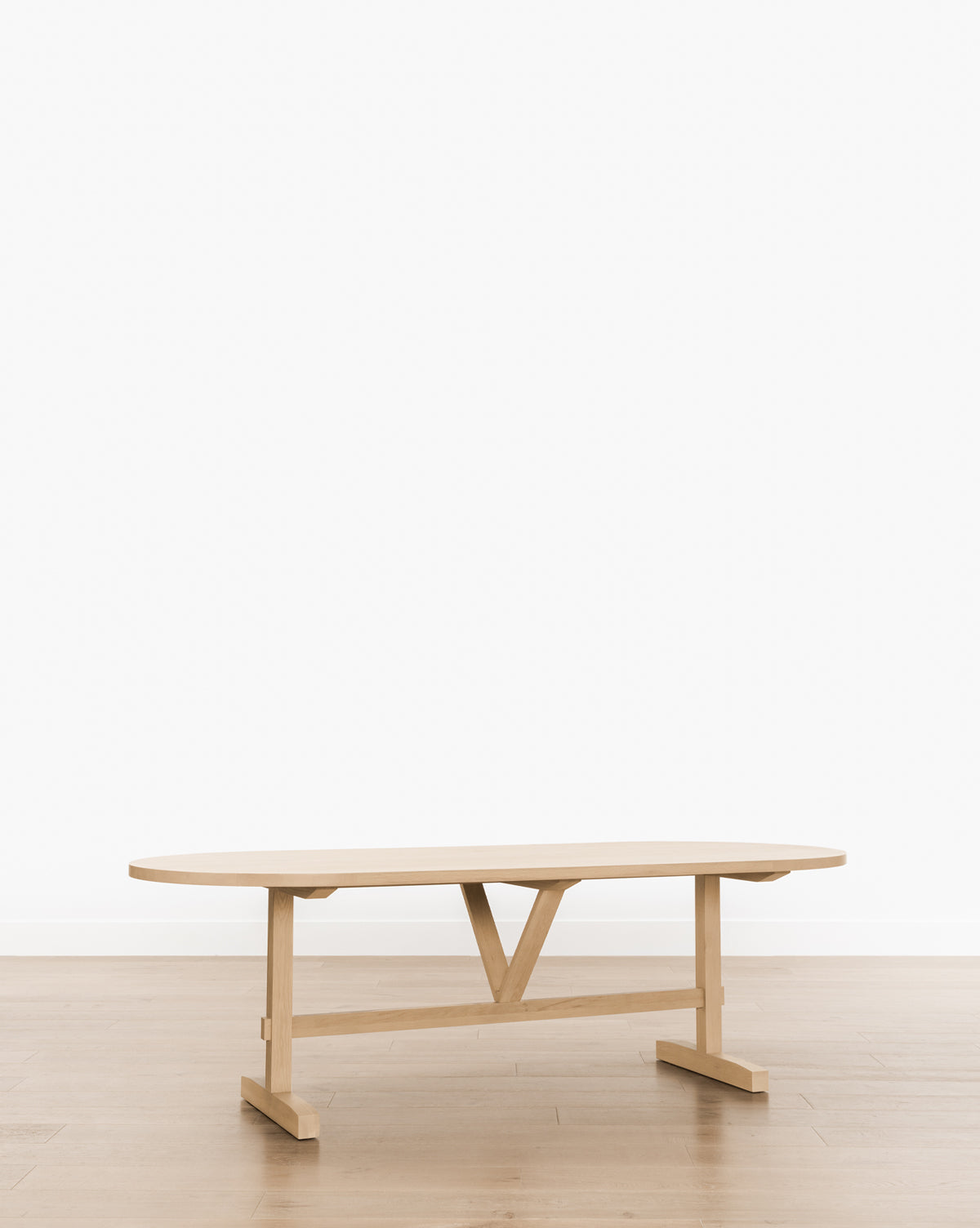 The Giselle Oval Dining Table, made of light wood with a distinctive V-shaped support, is set on a wooden floor against a plain white background by McGee & Co.