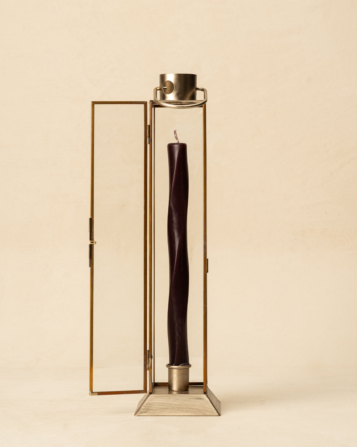 A tall, dark taper candle stands in the Glass & Metal Lantern Taper Holder with one door open, set against a plain beige background, by McGee & Co.